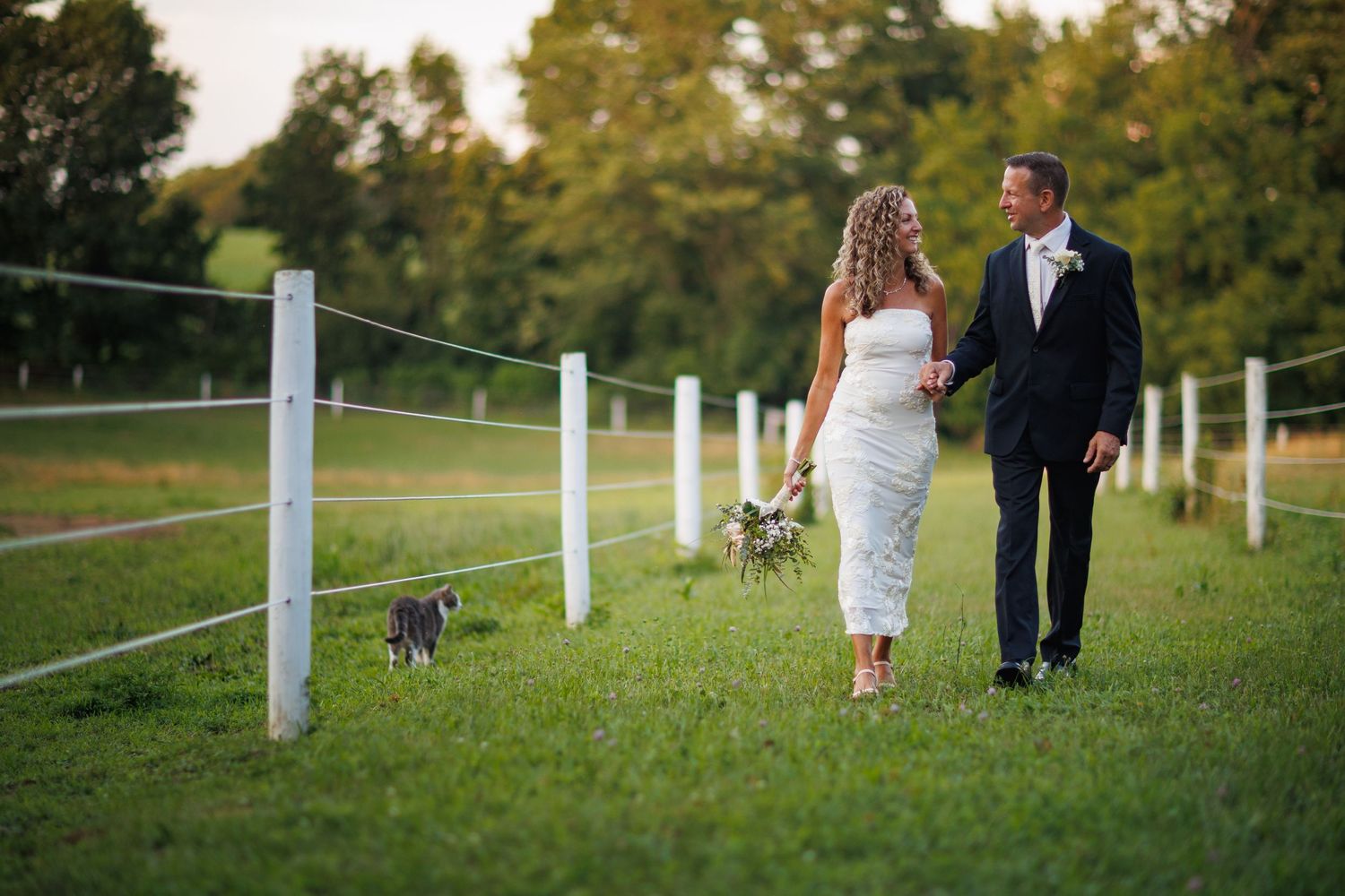 Bride and groom walk hand-in-hand along a white fence on a lush green farm, accompanied by a small dog.