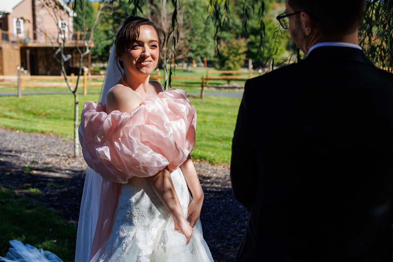 Bride in white wedding gown with pink floral wrap smiles at groom during outdoor ceremony in lush garden setting.