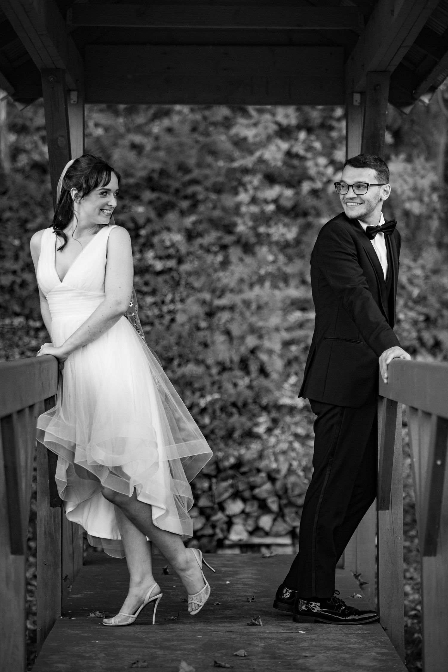 A smiling bride and groom pose on a wooden bridge surrounded by lush greenery.