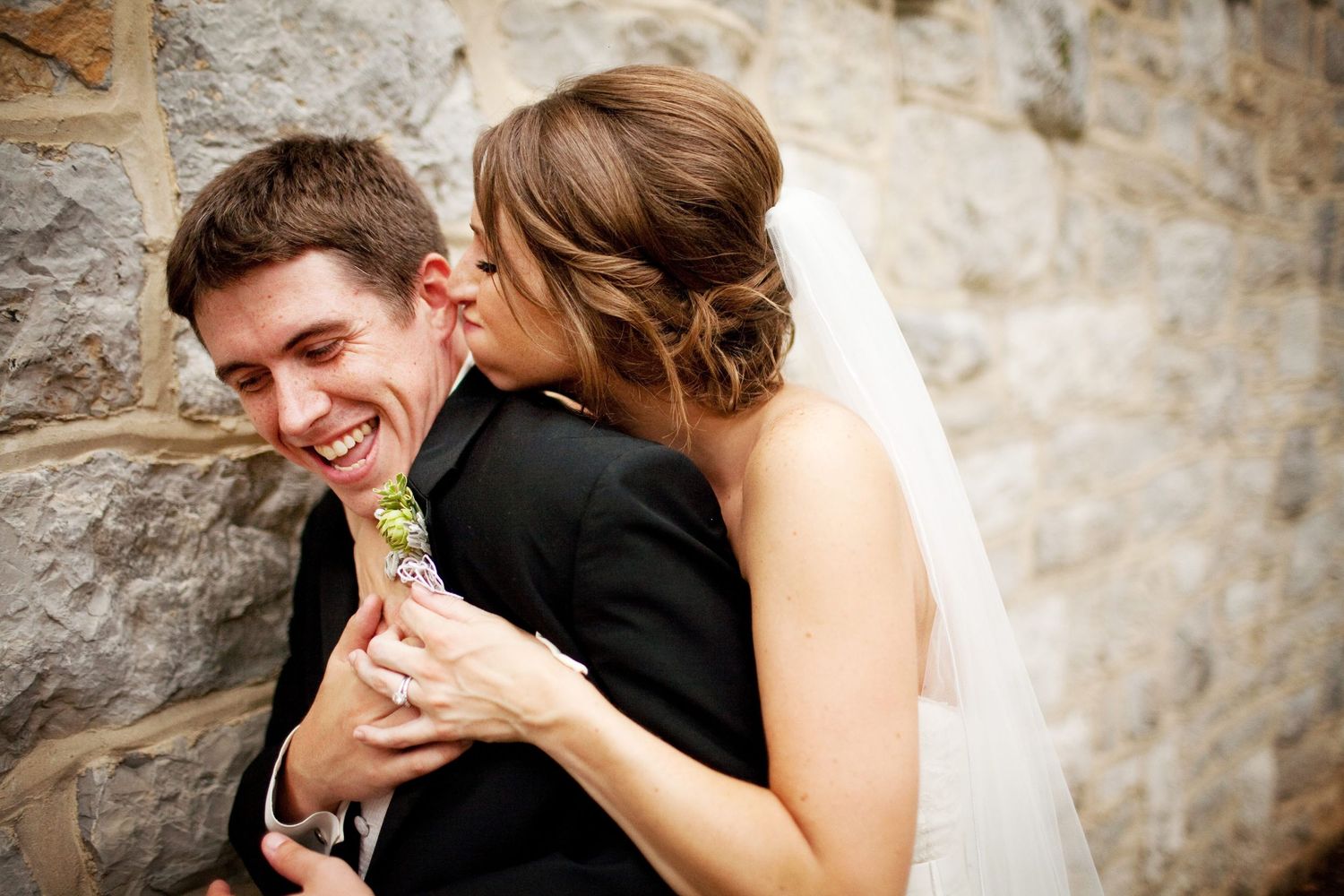 Bride kissing groom on cheek as he smiles against stone wall in wedding attire.