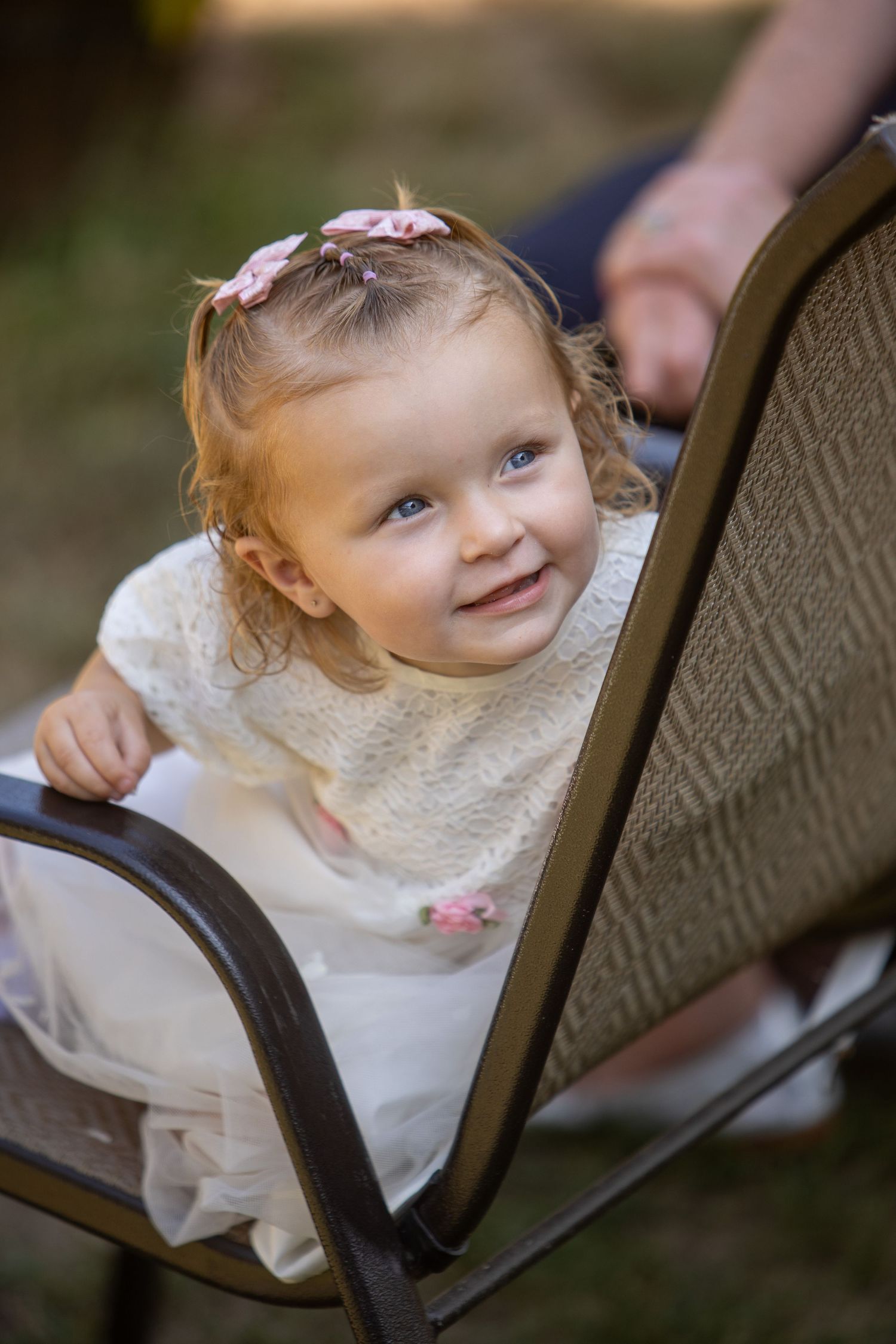 Young child in a white lace dress smiling while seated in a wicker chair outdoors.