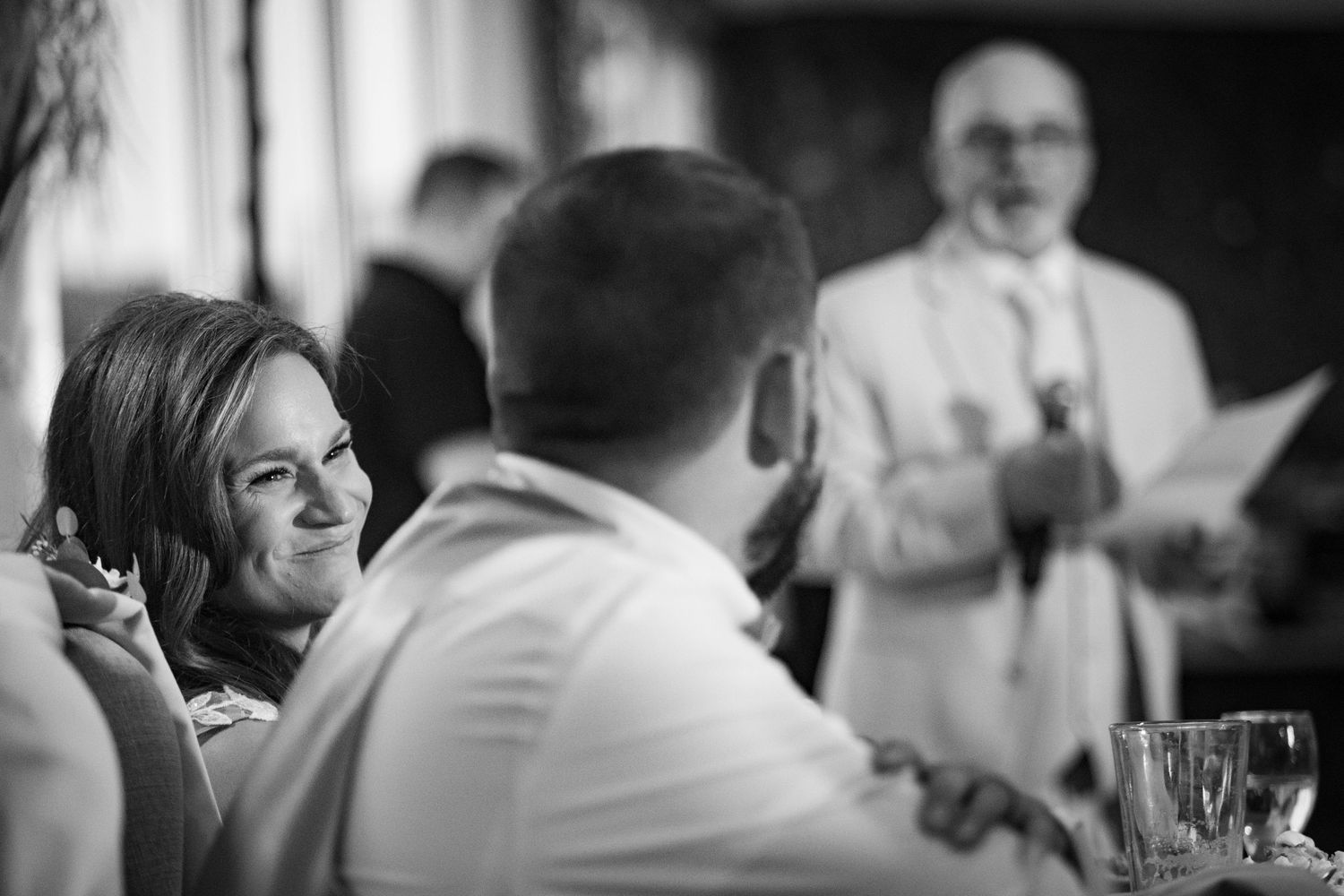 Black and white photo captures candid laughter during wedding reception toasts.