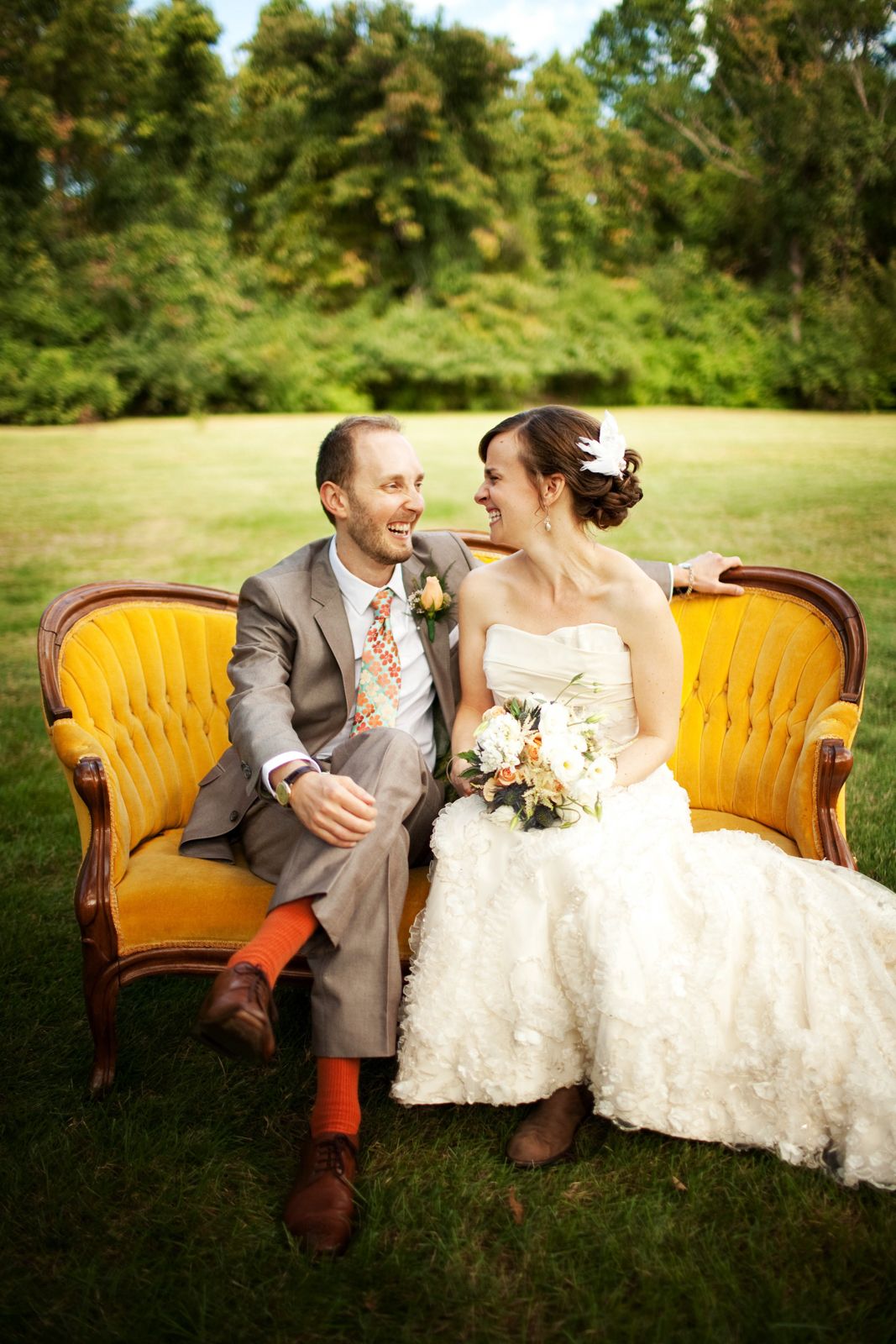 A couple shares joyful laughter while sitting together on a vintage yellow velvet sofa outdoors on a sunny day.