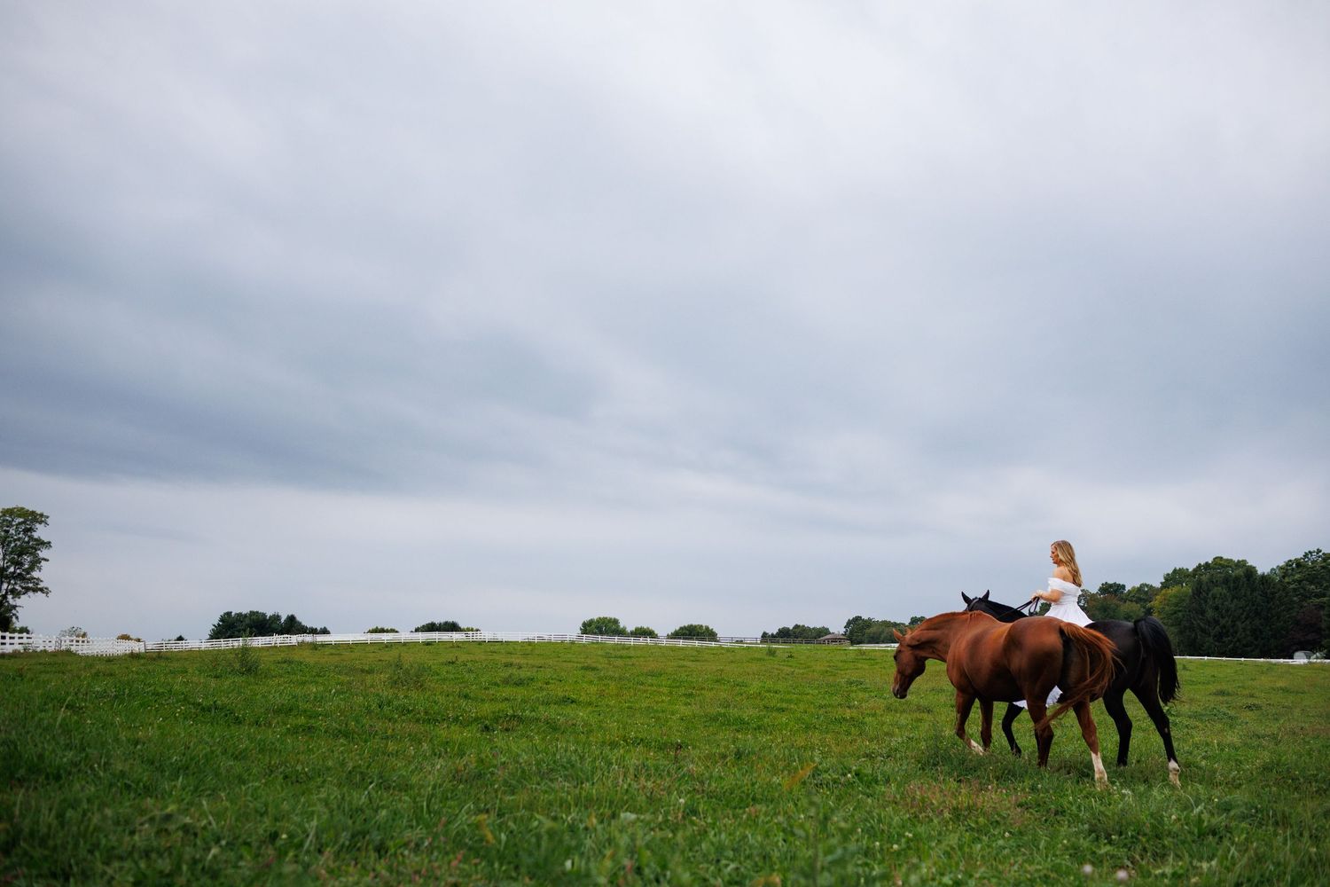 A woman rides a brown horse alongside a black horse across a lush green field under a cloudy sky.