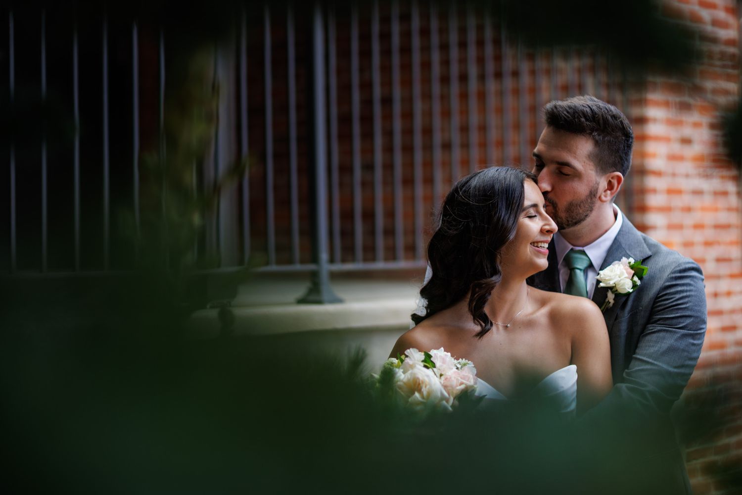 A groom in a gray suit kisses his smiling bride holding white flowers near a brick wall.