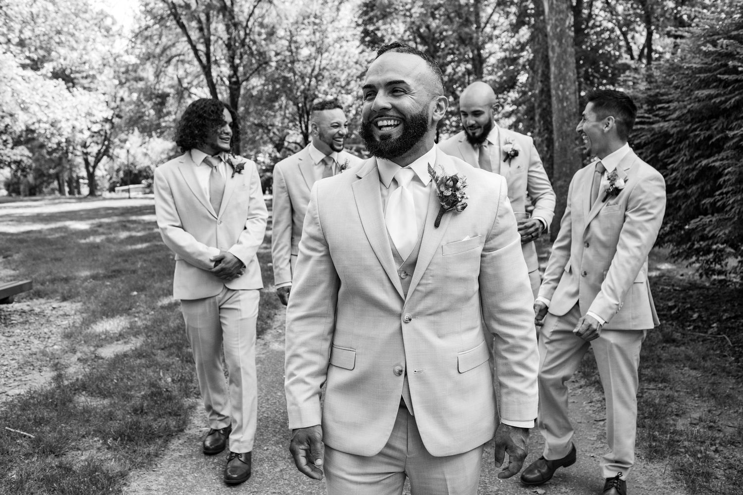 Groom smiling with groomsmen in tan suits on a tree-lined path at an outdoor wedding.