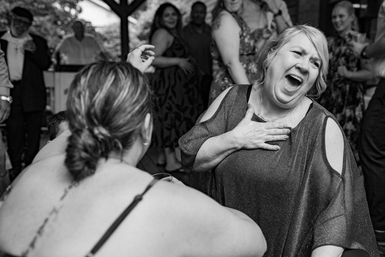 Black and white photo of a woman laughing with hand on chest while dancing at a wedding reception.