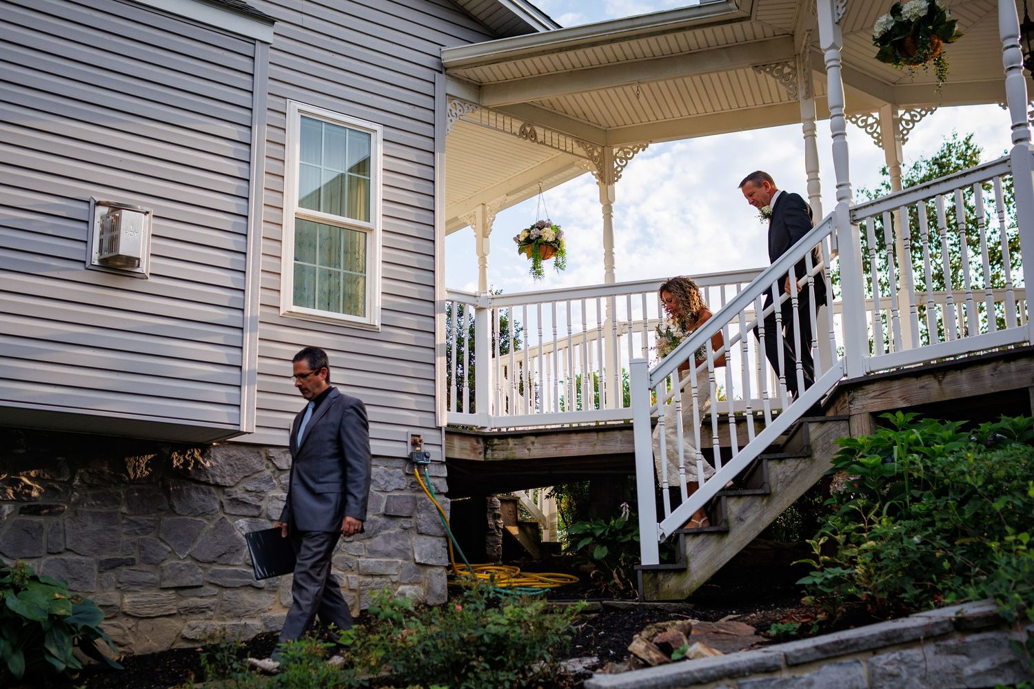 Elegant wedding party descends white porch staircase of a charming gray clapboard house with decorative railings.