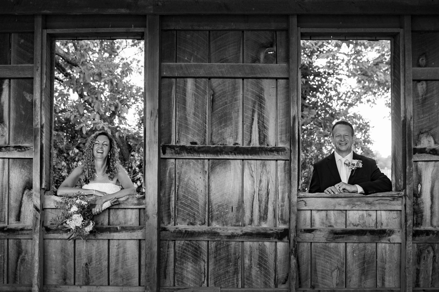 Black and white photo of bride and groom leaning out of rustic wooden barn windows on their wedding day.