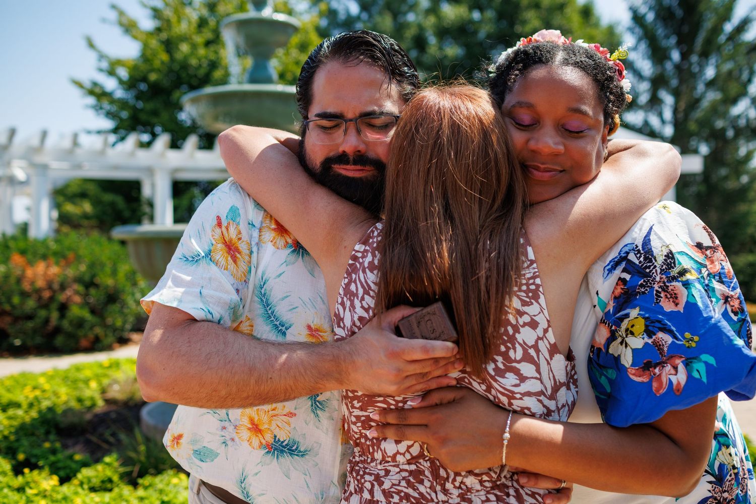 Three people sharing an emotional group hug outdoors near a fountain on a sunny day.