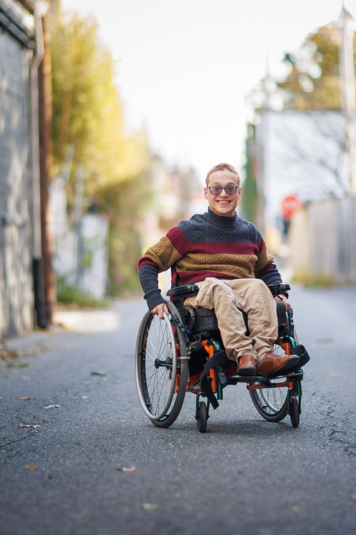 Person in a wheelchair wearing a striped sweater and sunglasses poses outdoors on a fall day with autumn leaves on ground.