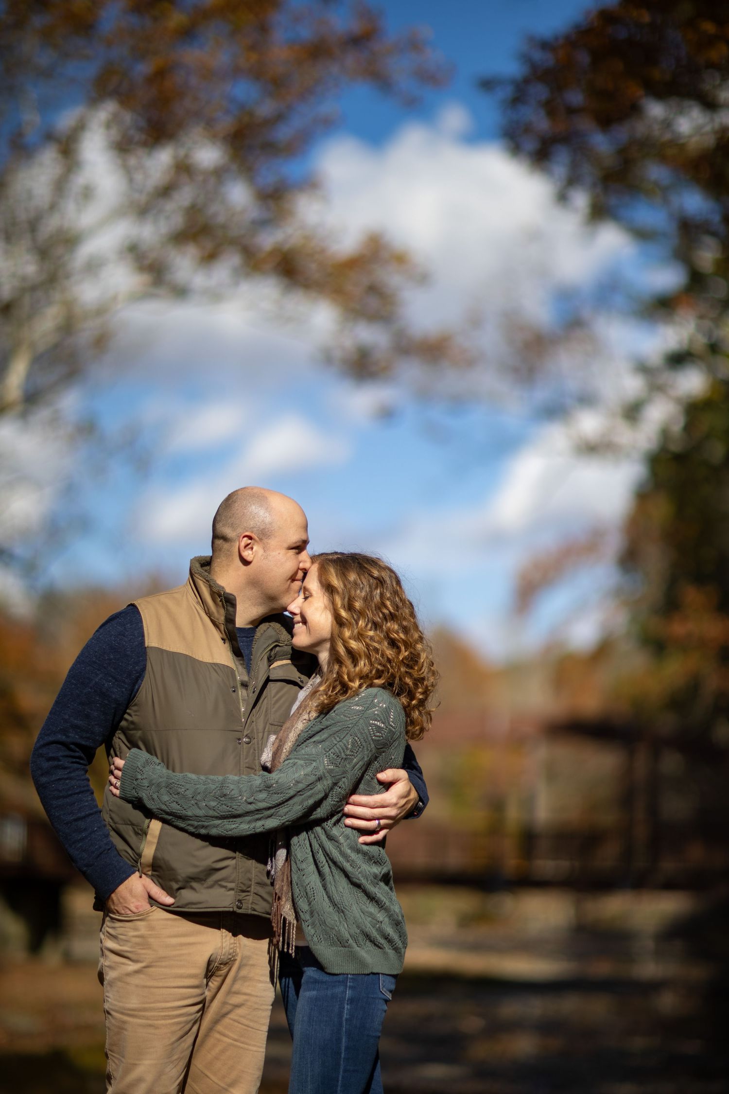 A romantic autumn moment shared between a couple embracing outdoors with fall foliage and blue sky in the background.