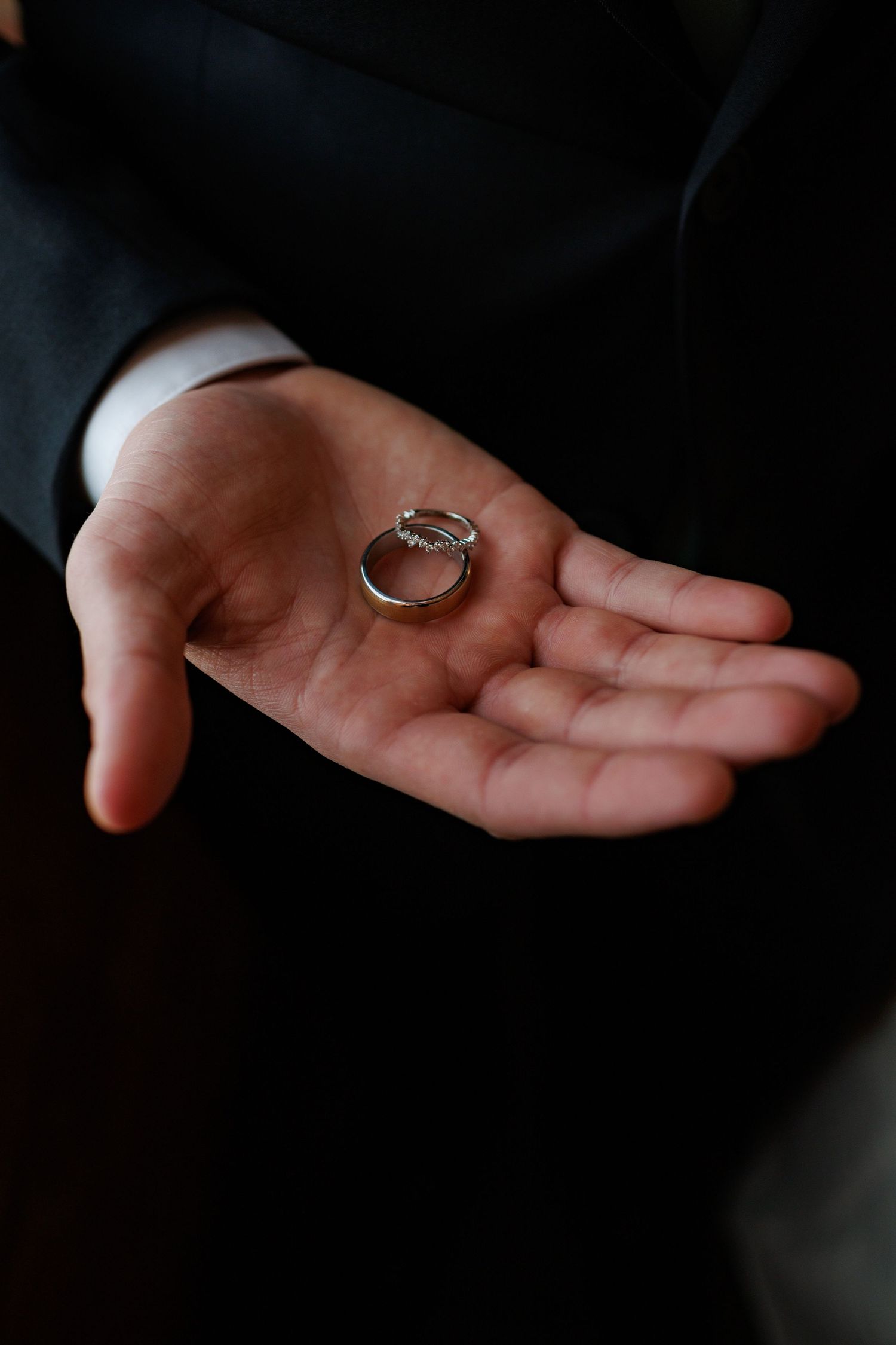 Groom in black suit holding two wedding rings in his open palm.