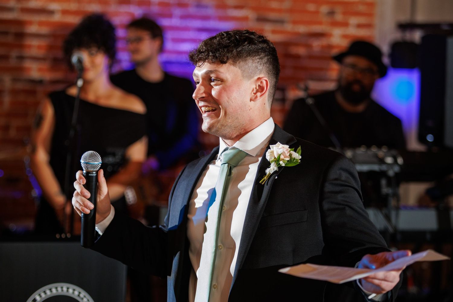 Smiling groomsman delivers wedding speech holding microphone and notes with live band performing behind him.
