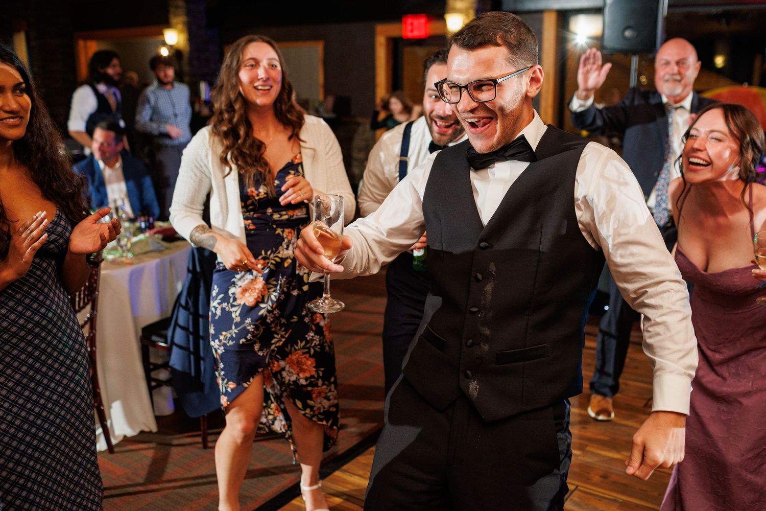 A man in a bow tie and vest dances enthusiastically surrounded by laughing wedding guests.