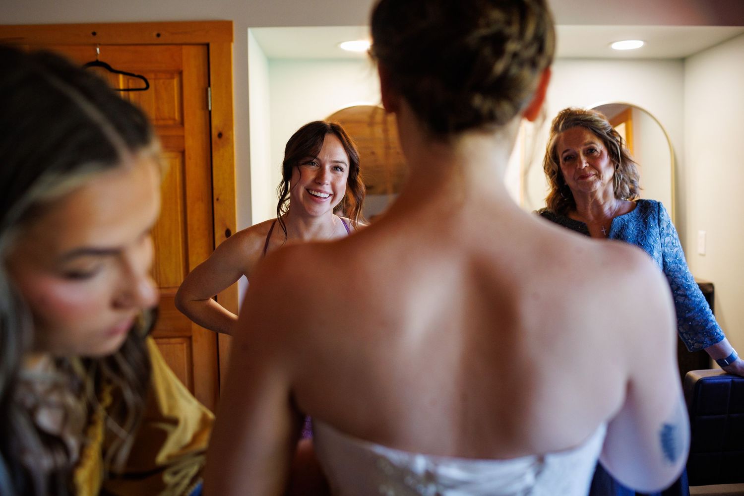 Bride getting dressed with smiling bridesmaids and mother helping in a warm wooden room.