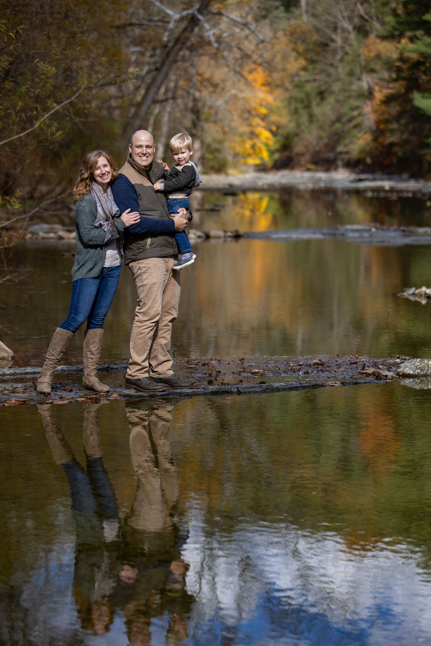 Couple standing on fallen log over tranquil pond with autumn foliage reflecting in calm water.