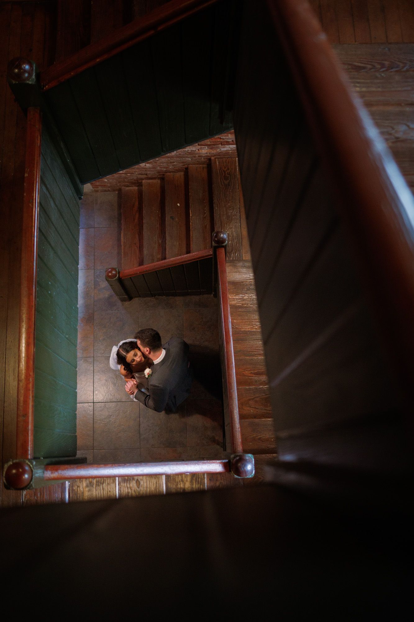 Aerial view of couple embracing at the bottom of a wooden staircase in a dimly lit venue.
