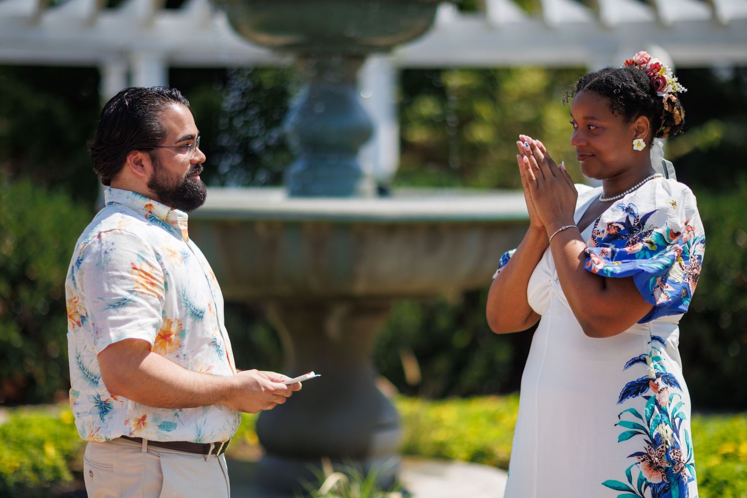 A man and woman exchange vows outdoors near a fountain, with the woman holding her hands in prayer.
