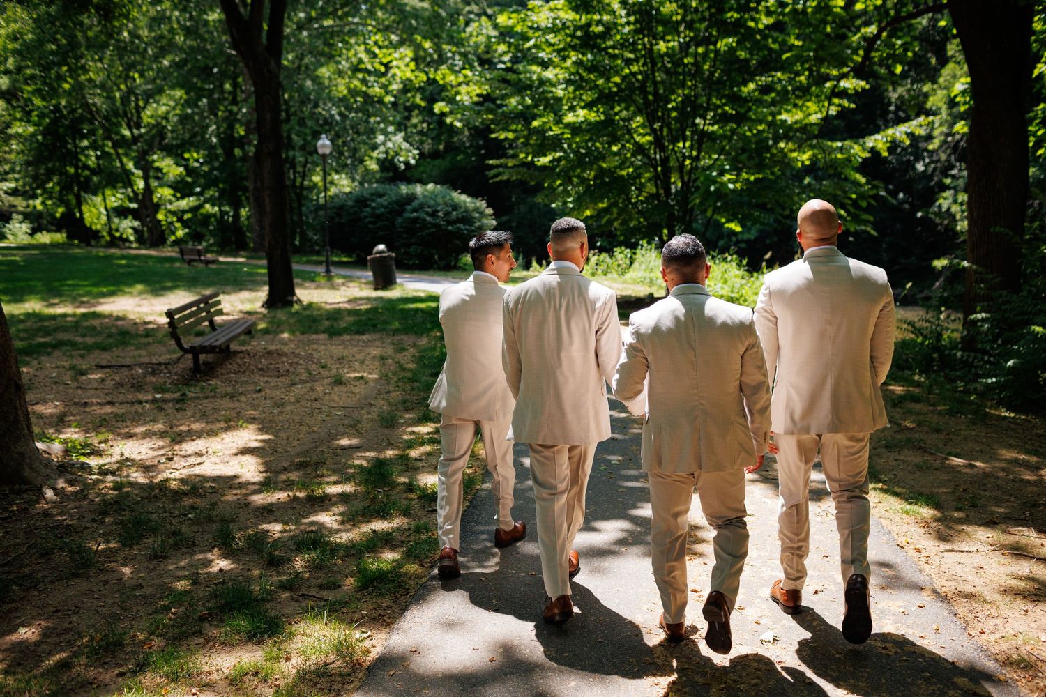 Four groomsmen in light suits walking together on a sunny park path surrounded by lush green trees.