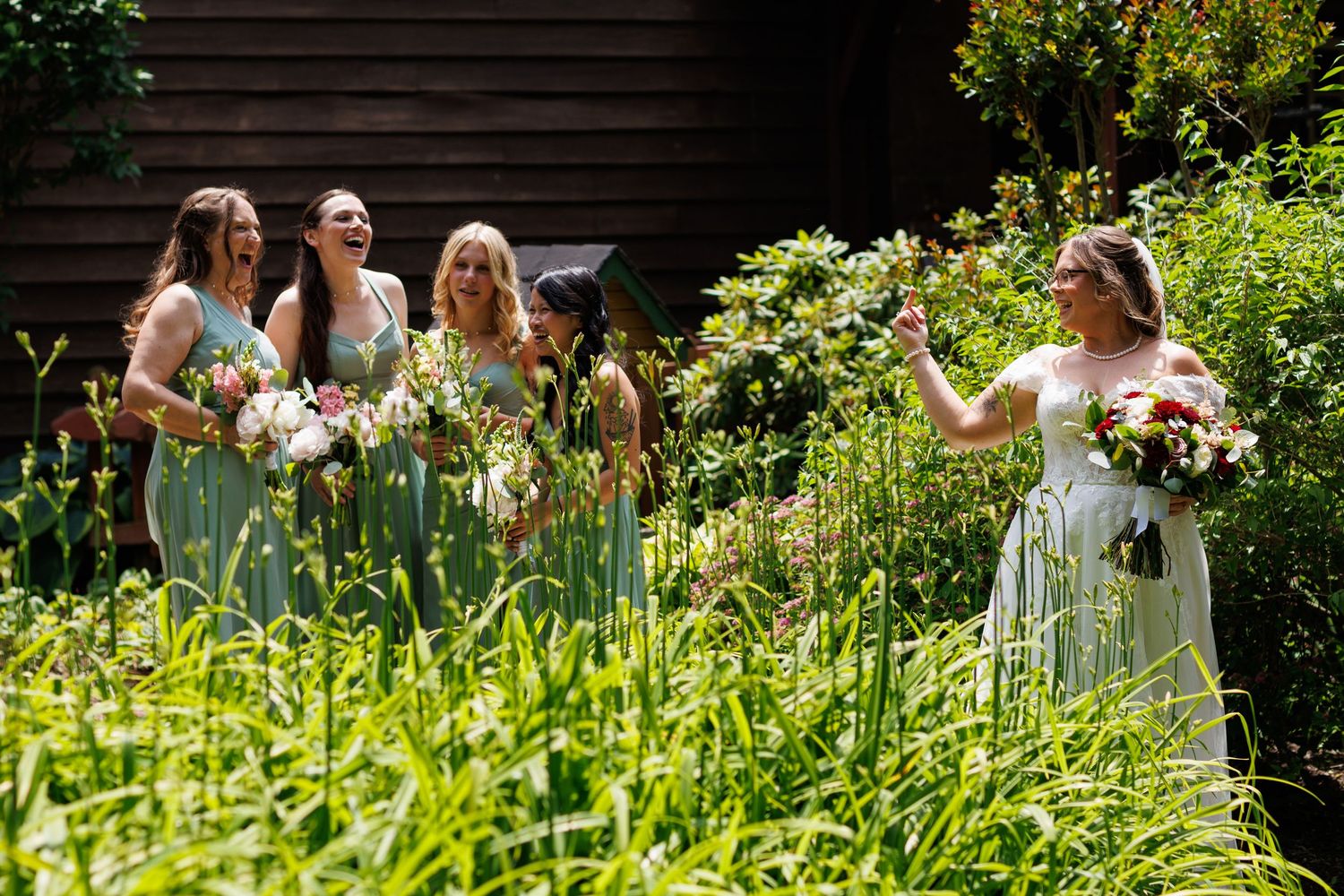 Bride and bridesmaids in sage green dresses laughing together in a lush garden setting.