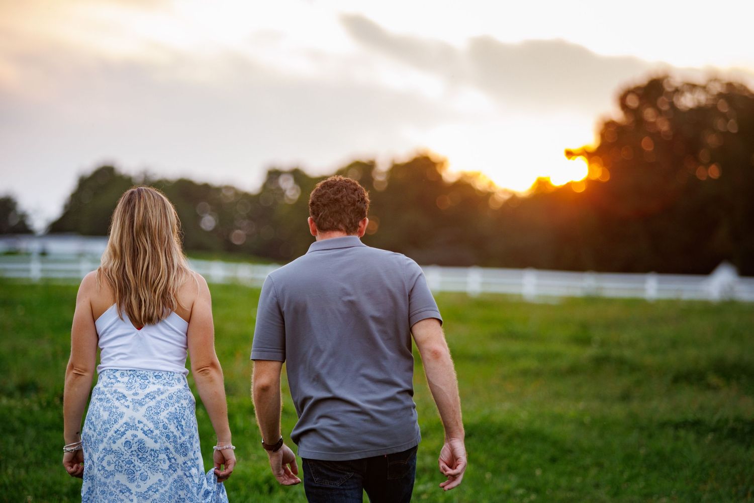 Couple walking hand in hand through a green field toward a beautiful sunset near a white fence.