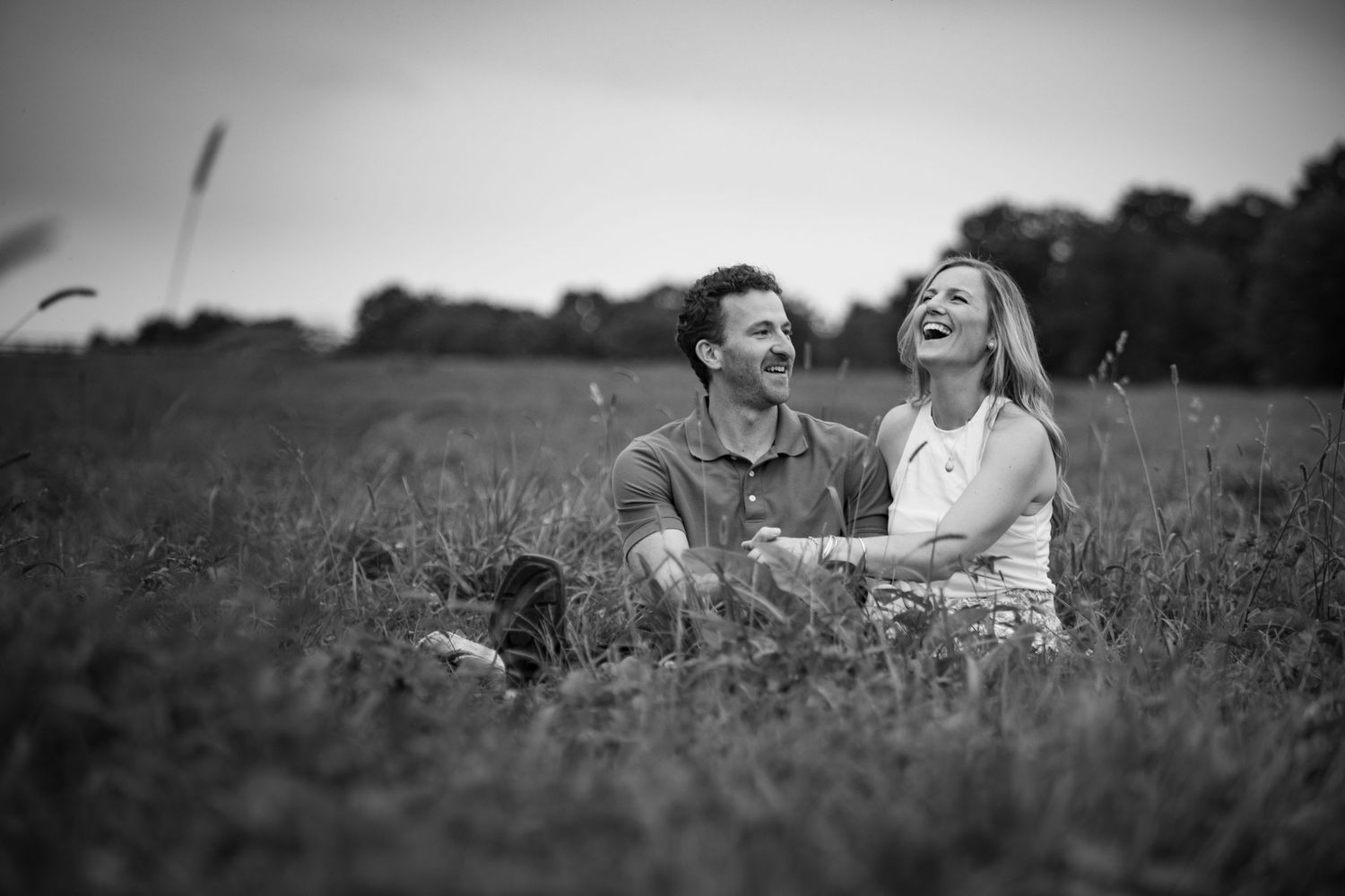 Black and white photo of a laughing couple sitting together in a grassy field at dusk.