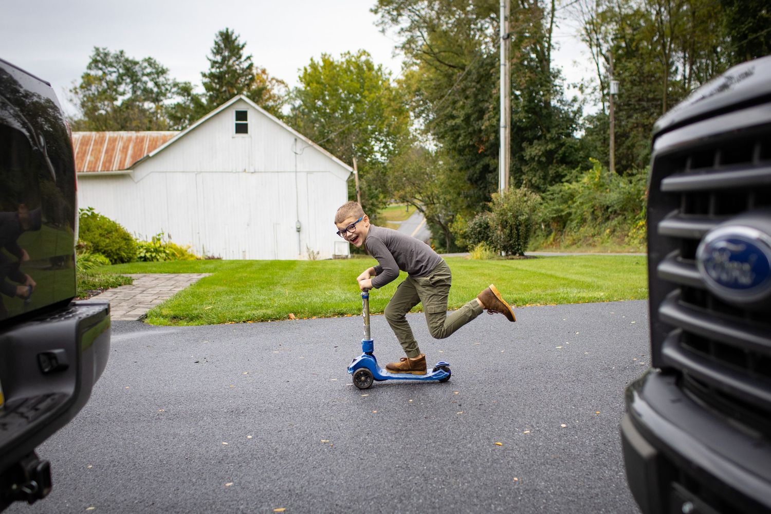 Person riding a blue scooter on a residential street with parked cars and trees in the background.