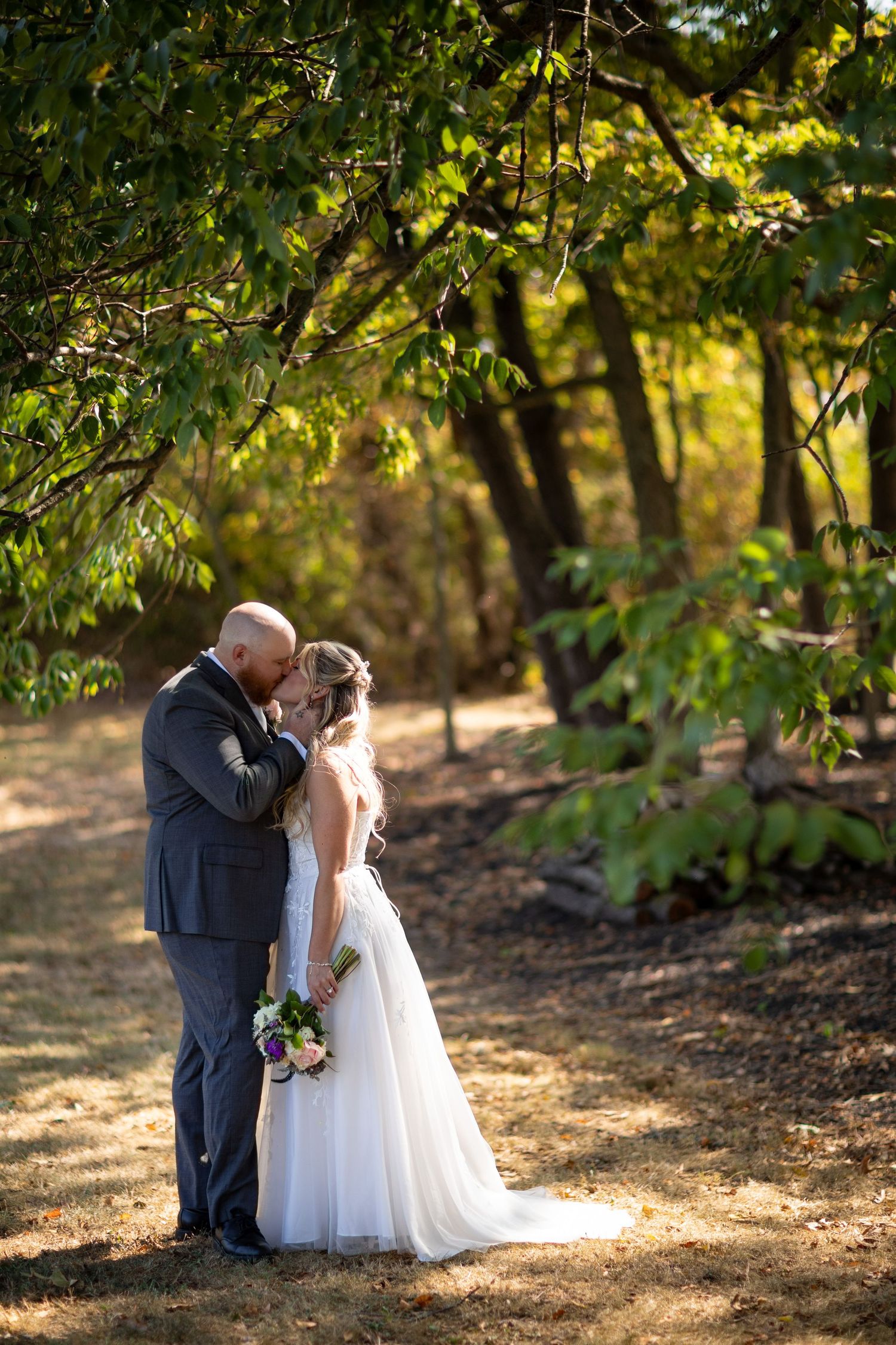 A newlywed couple shares an intimate moment in a sunlit wooded area during their wedding photo session.