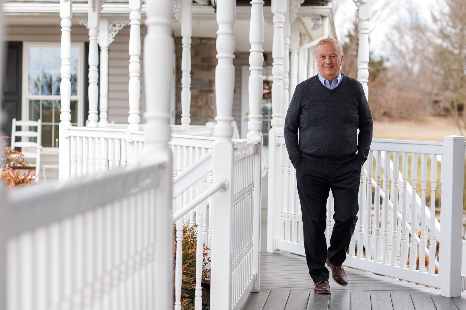 A person stands on a white wraparound porch wearing a black sweater and dark pants on a winter day.