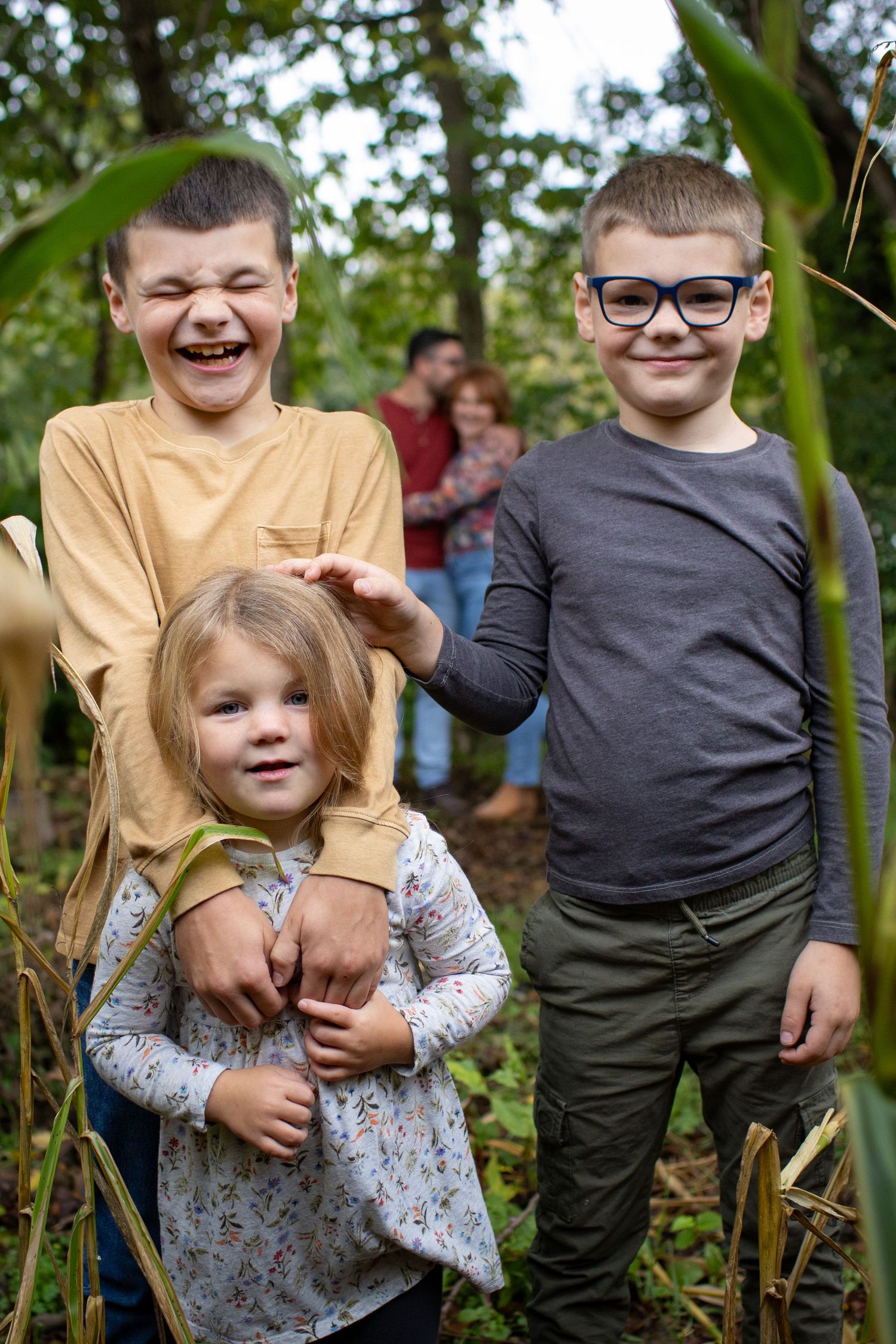 Three children share joyful moments together while standing among tall plants in a natural outdoor setting.