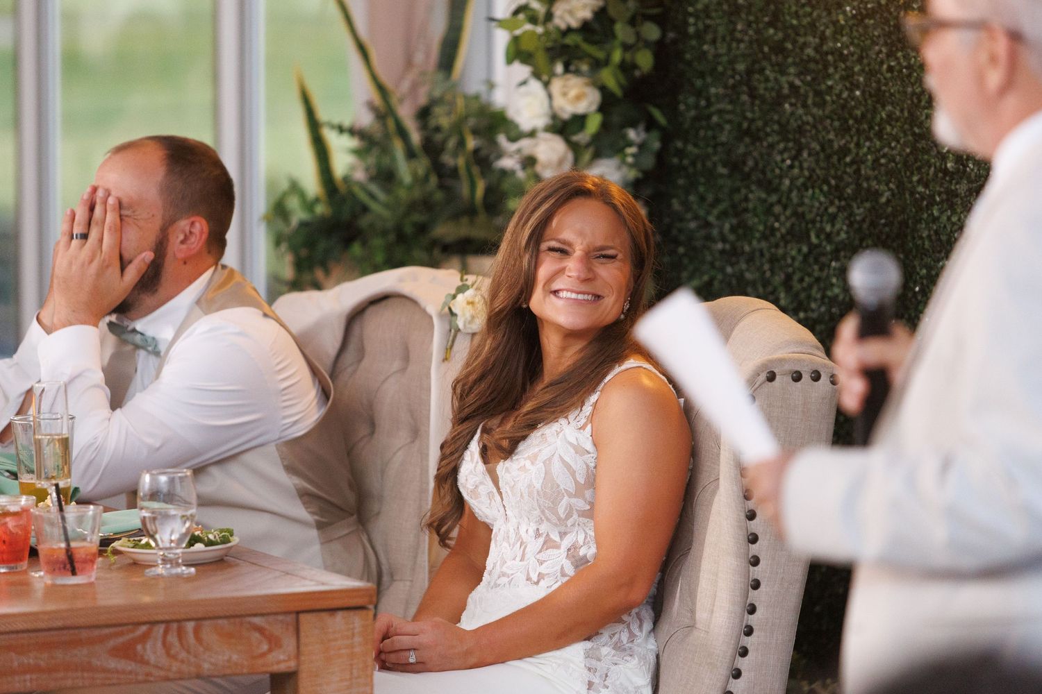 Wedding guests laugh and enjoy themselves at an elegant reception with upholstered chairs and greenery backdrop.