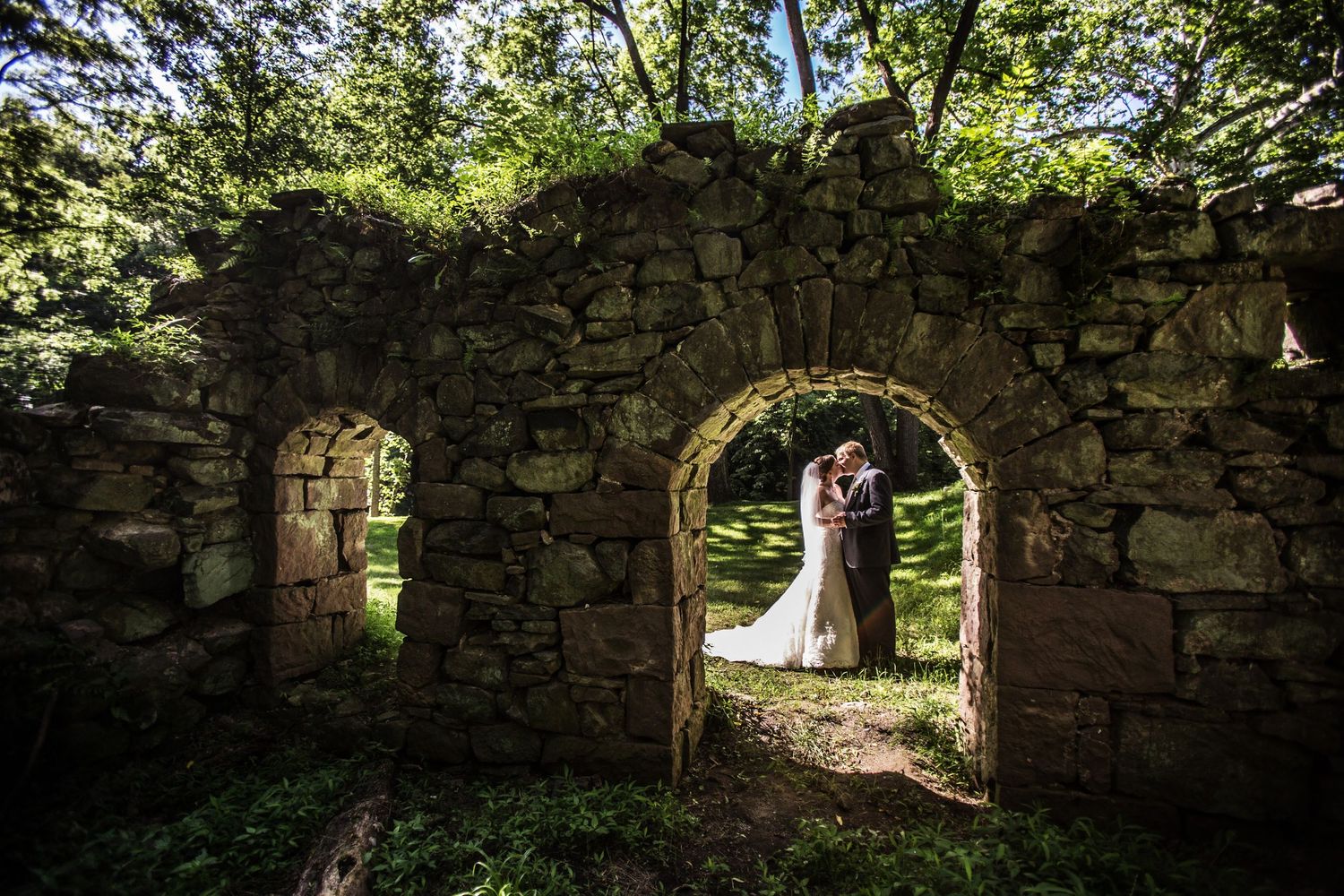 Wedding couple standing in stone archway ruins surrounded by lush green forest.