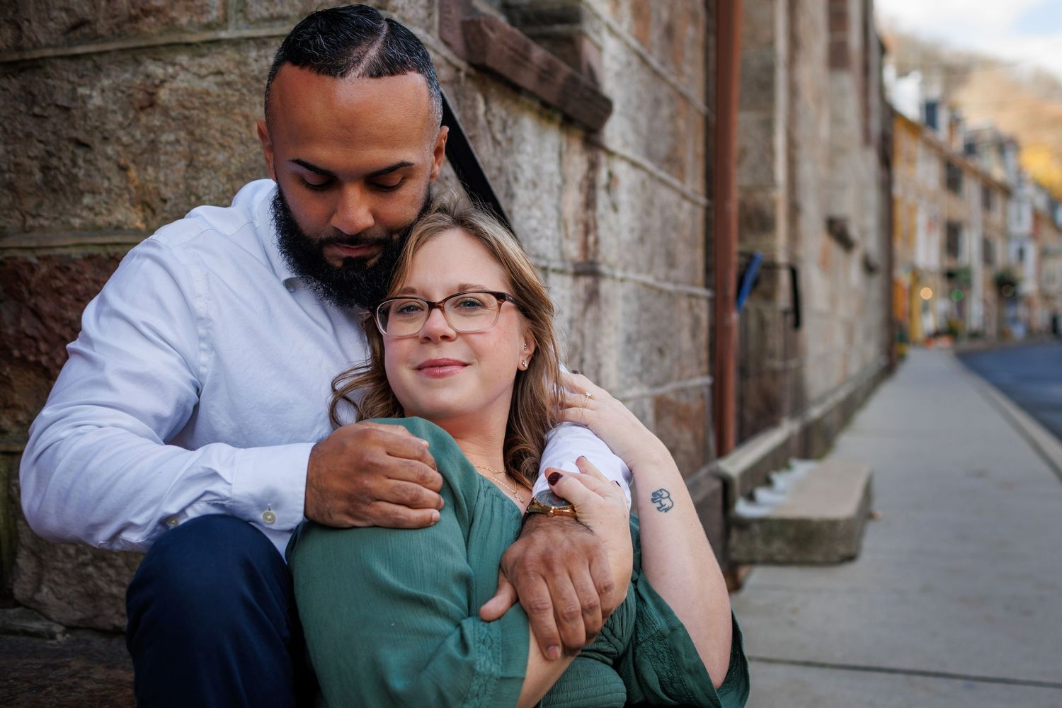 A couple shares an intimate embrace against a rustic stone wall in a European city street.