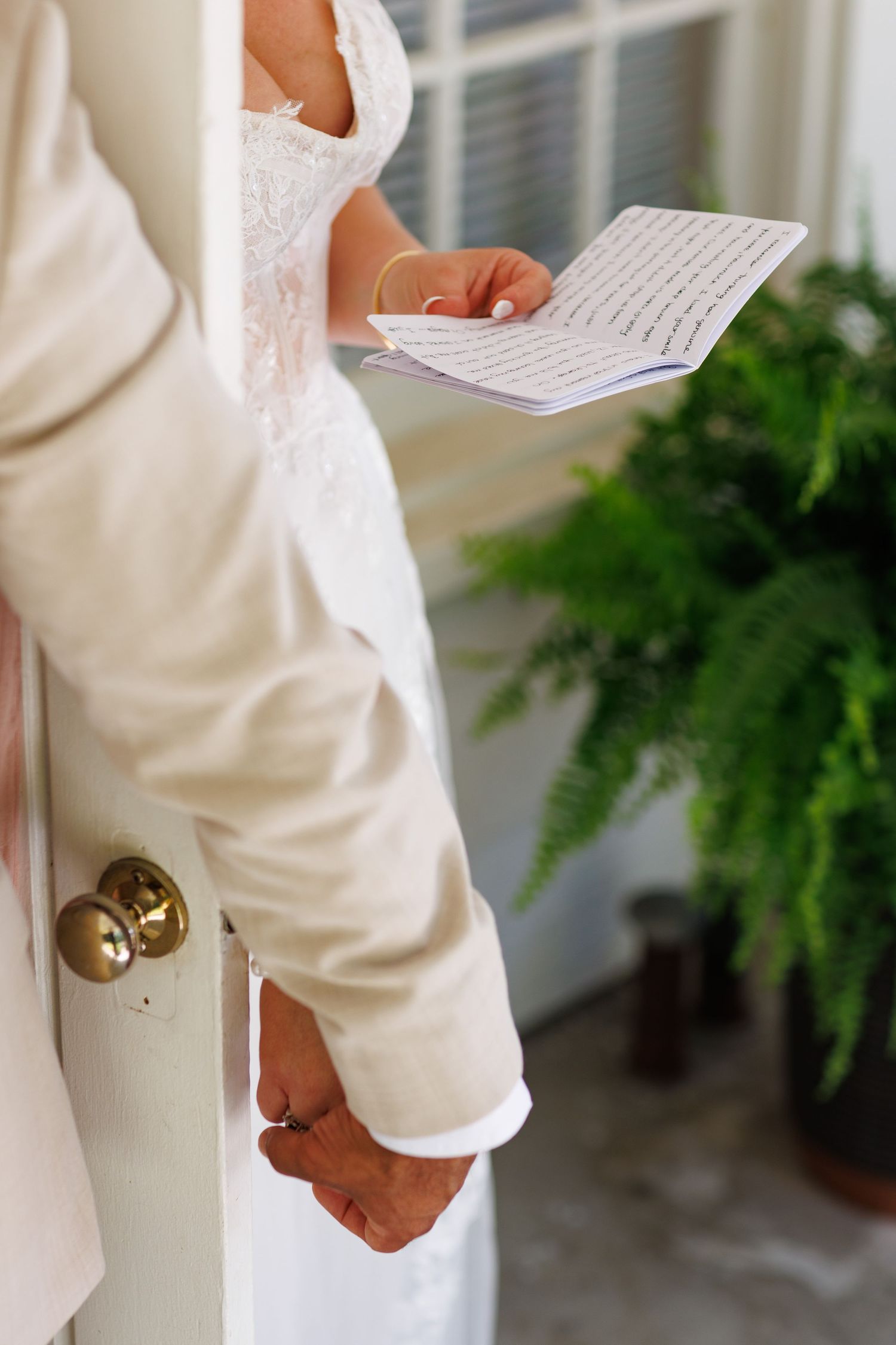 Bride and groom in white attire holding hands and reading vows from a small notebook at a doorway.