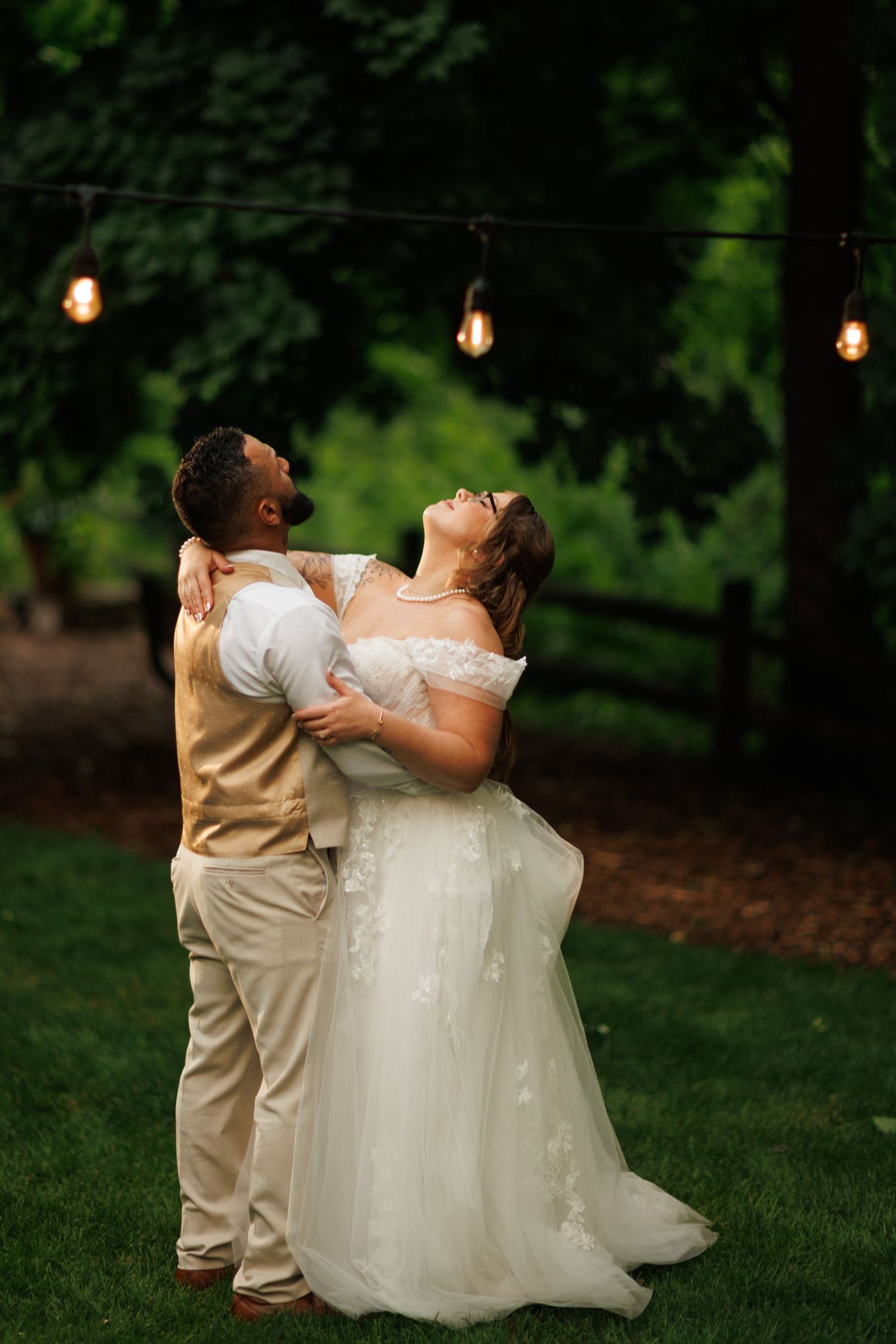 Bride and groom share romantic first dance under string lights in lush green garden at night.