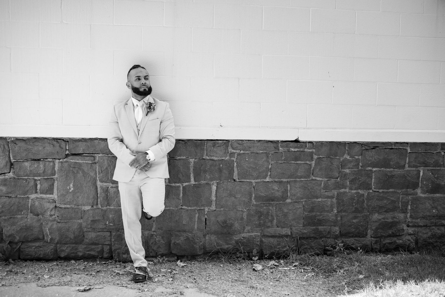 Groom in tan suit leaning against stone wall in black and white wedding portrait.