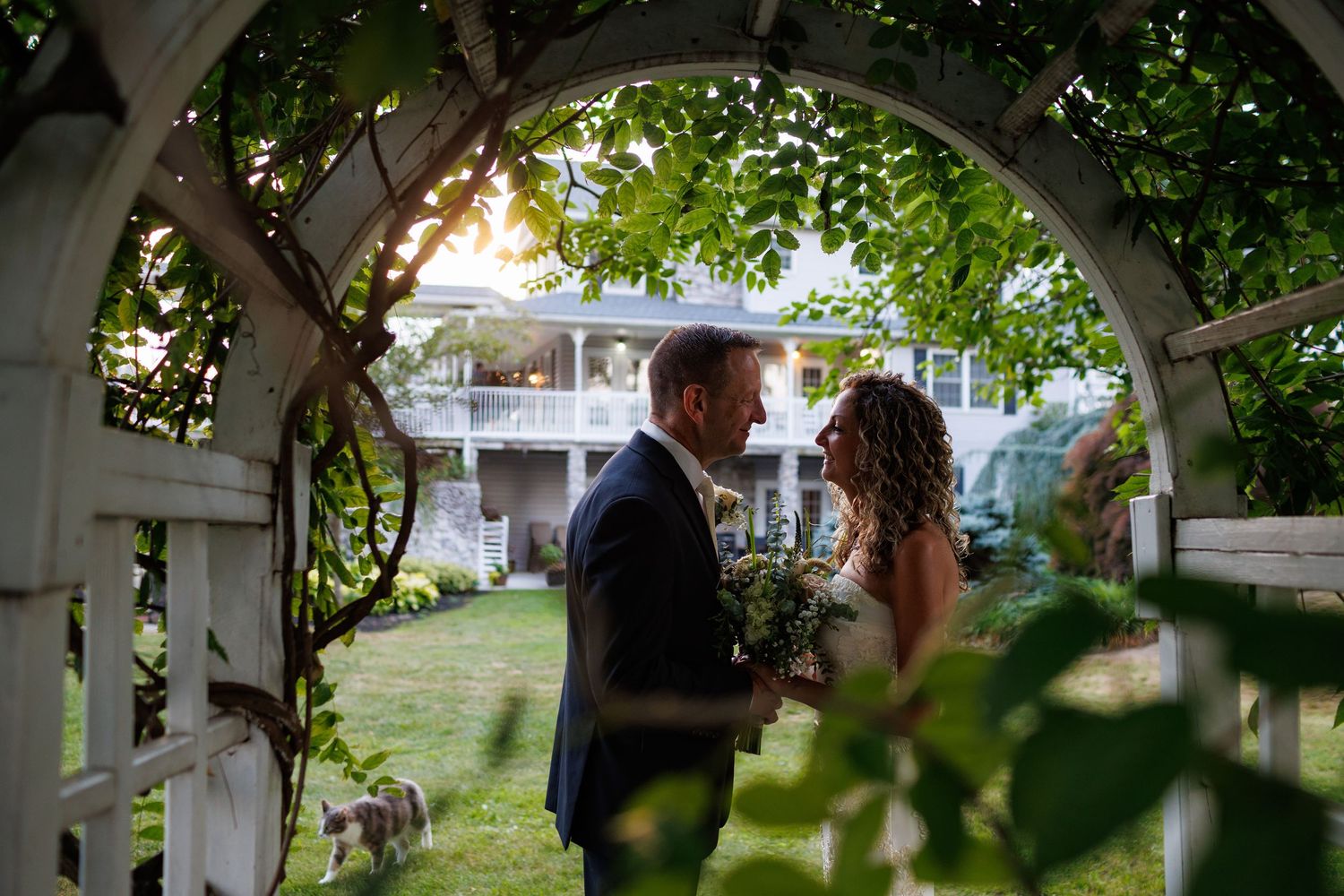 Bride and groom share a romantic moment framed by a white garden arbor with lush greenery at a wedding venue.