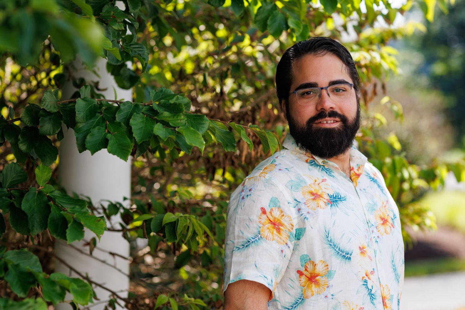 A bearded man wearing glasses and a floral shirt smiles outdoors beside lush green foliage.