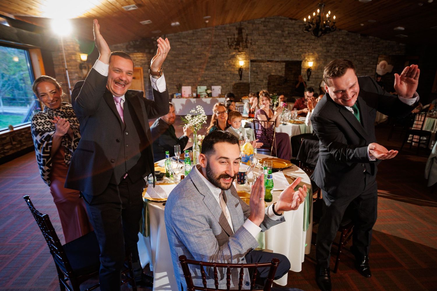 Joyful wedding reception guests raise hands and celebrate at elegantly decorated dinner tables.