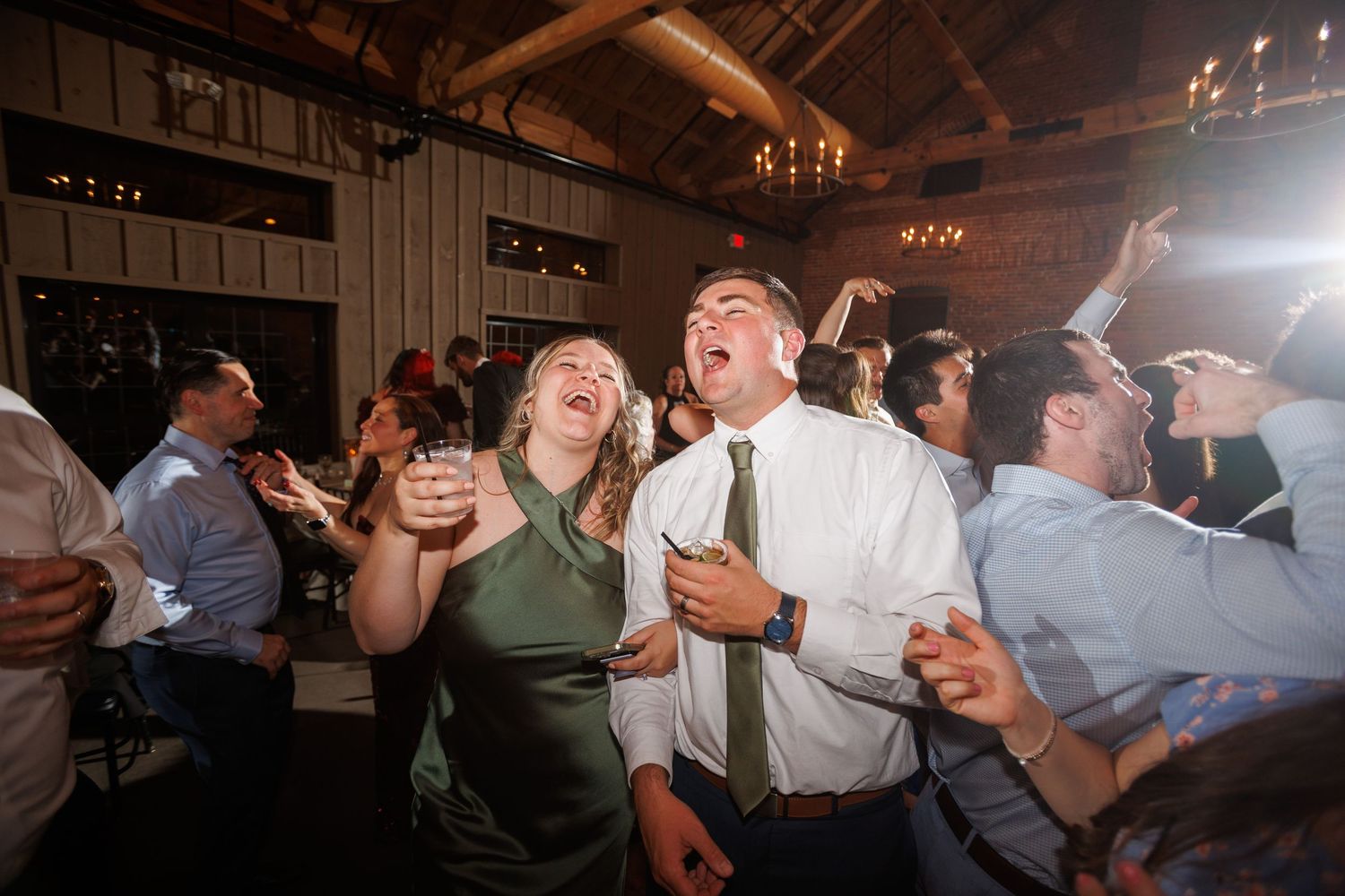 Wedding guests laugh and dance energetically on the dance floor during a lively reception celebration.