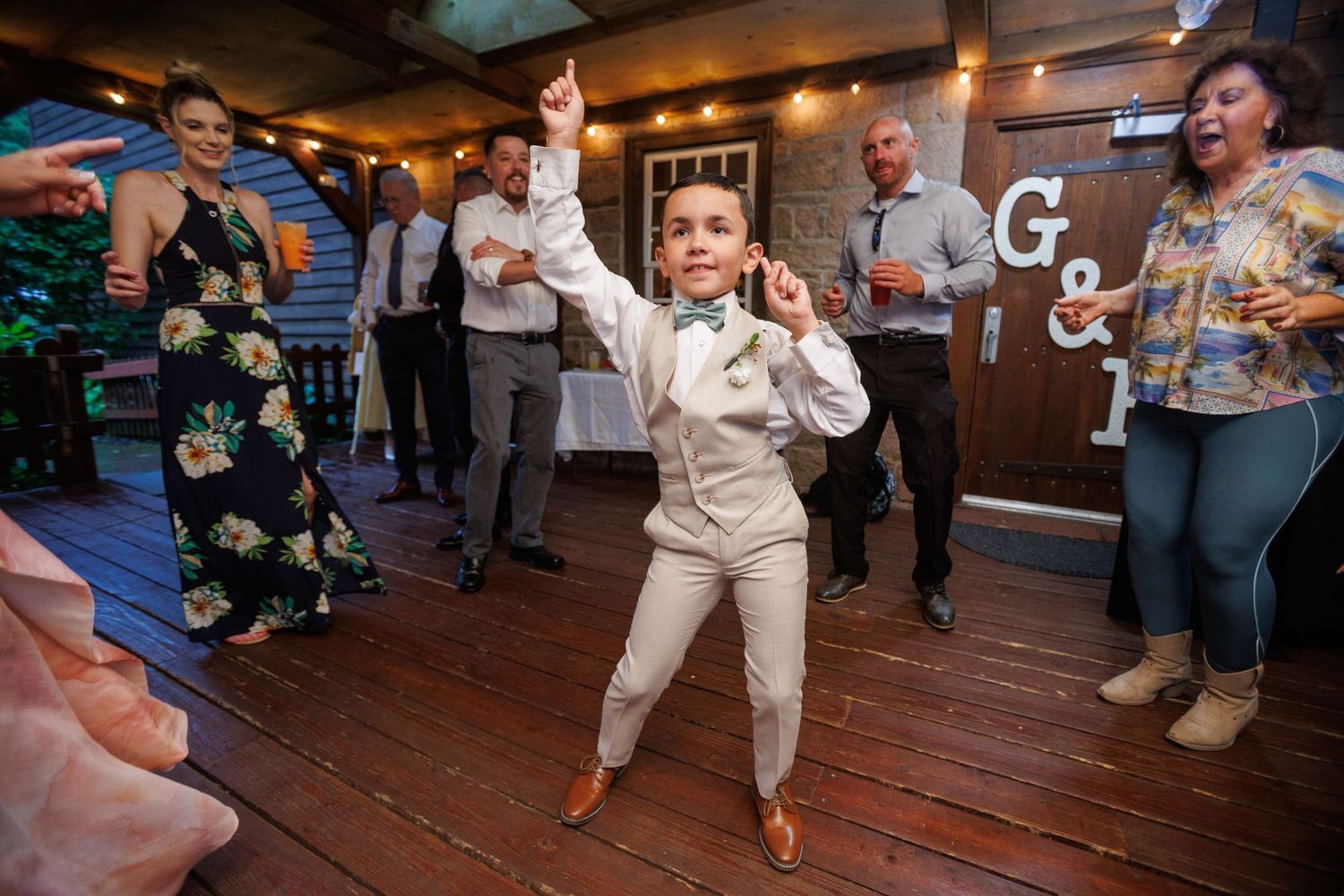 Energetic young boy in tan suit dancing joyfully at outdoor wedding reception celebration.