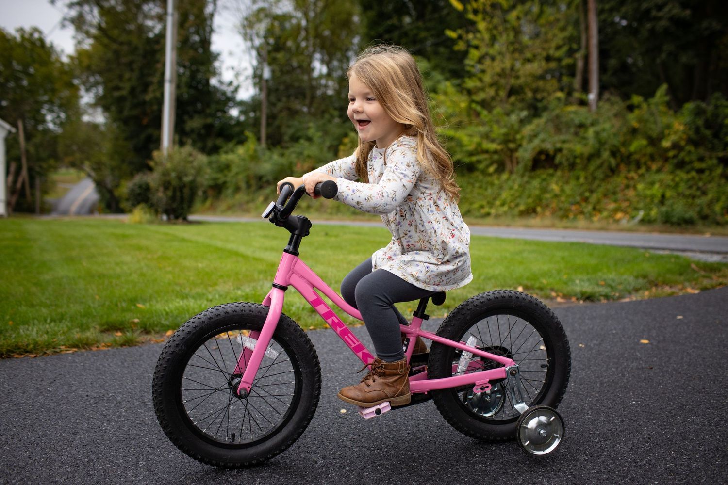 Pink children's balance bike parked on an asphalt path surrounded by green grass and trees.