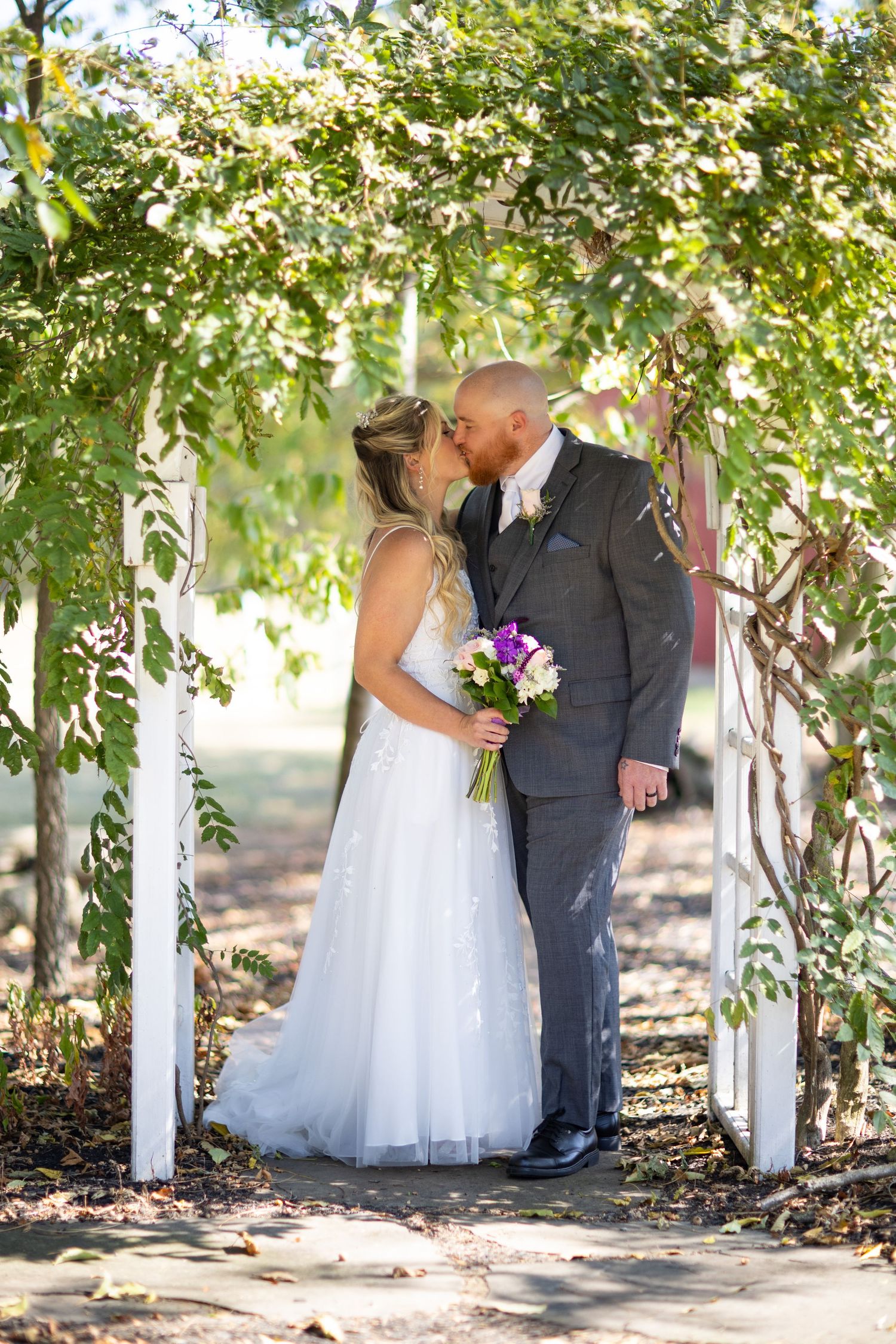 A romantic wedding couple kisses under a floral garden arch decorated with white draping and greenery.
