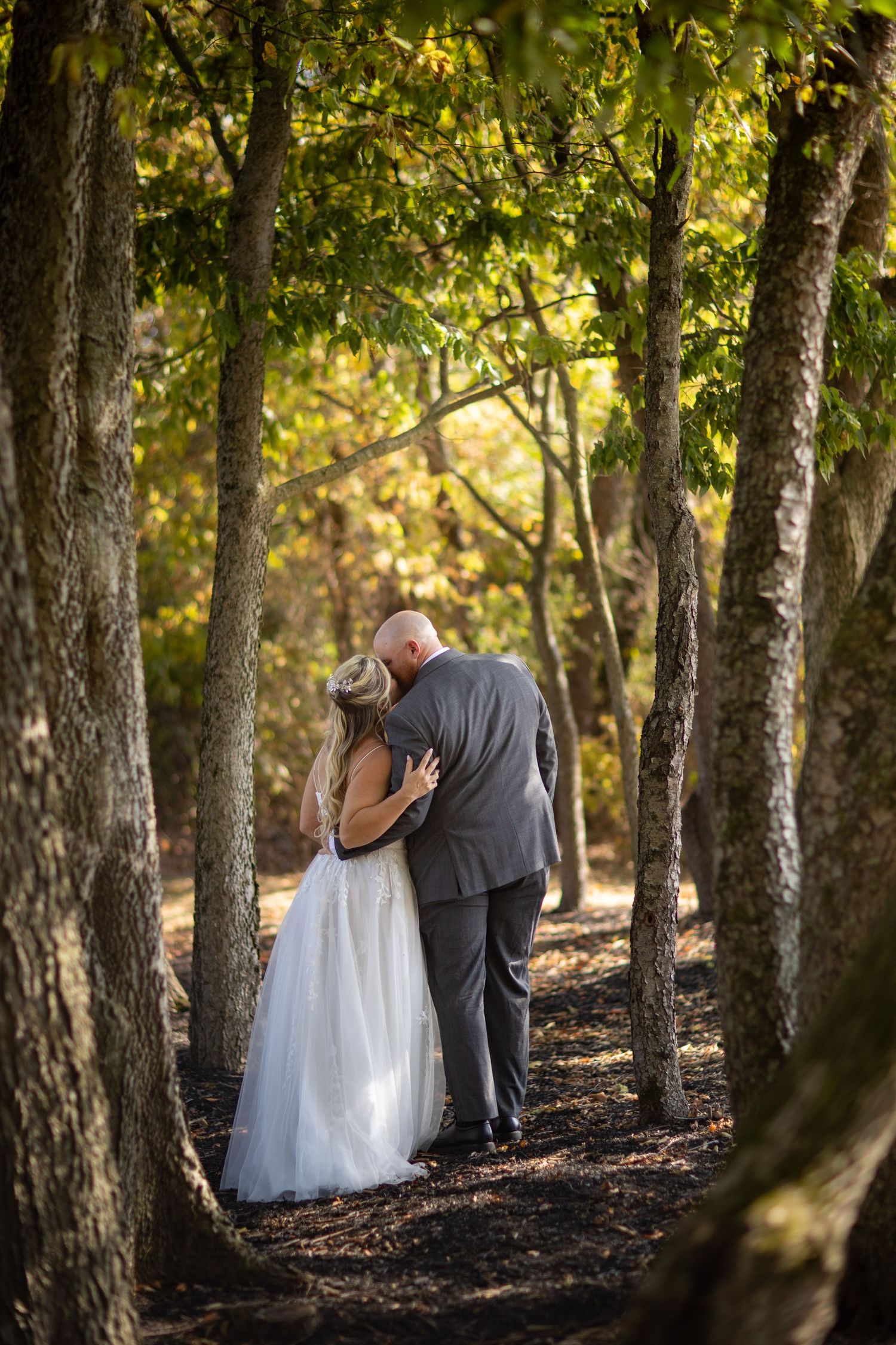 A couple shares an intimate moment walking together on a wooded path in autumn sunlight.