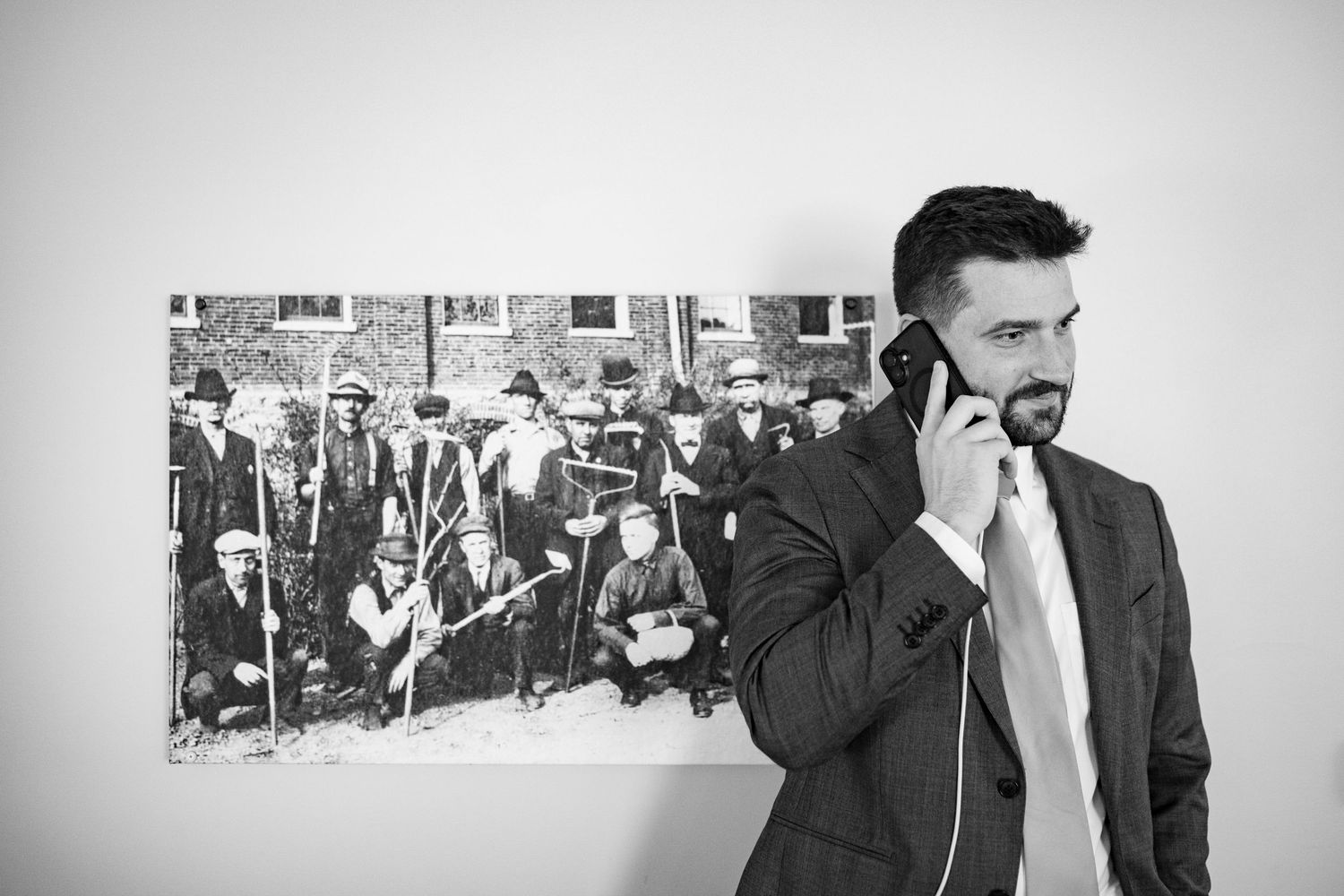 Black and white photo of a bearded man in a suit on a phone call, standing beside a vintage group portrait.