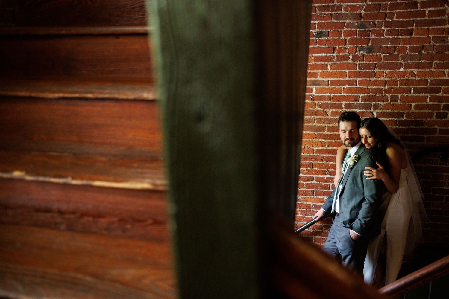 Bride and groom share intimate moment on staircase framed by warm wooden and exposed brick walls.