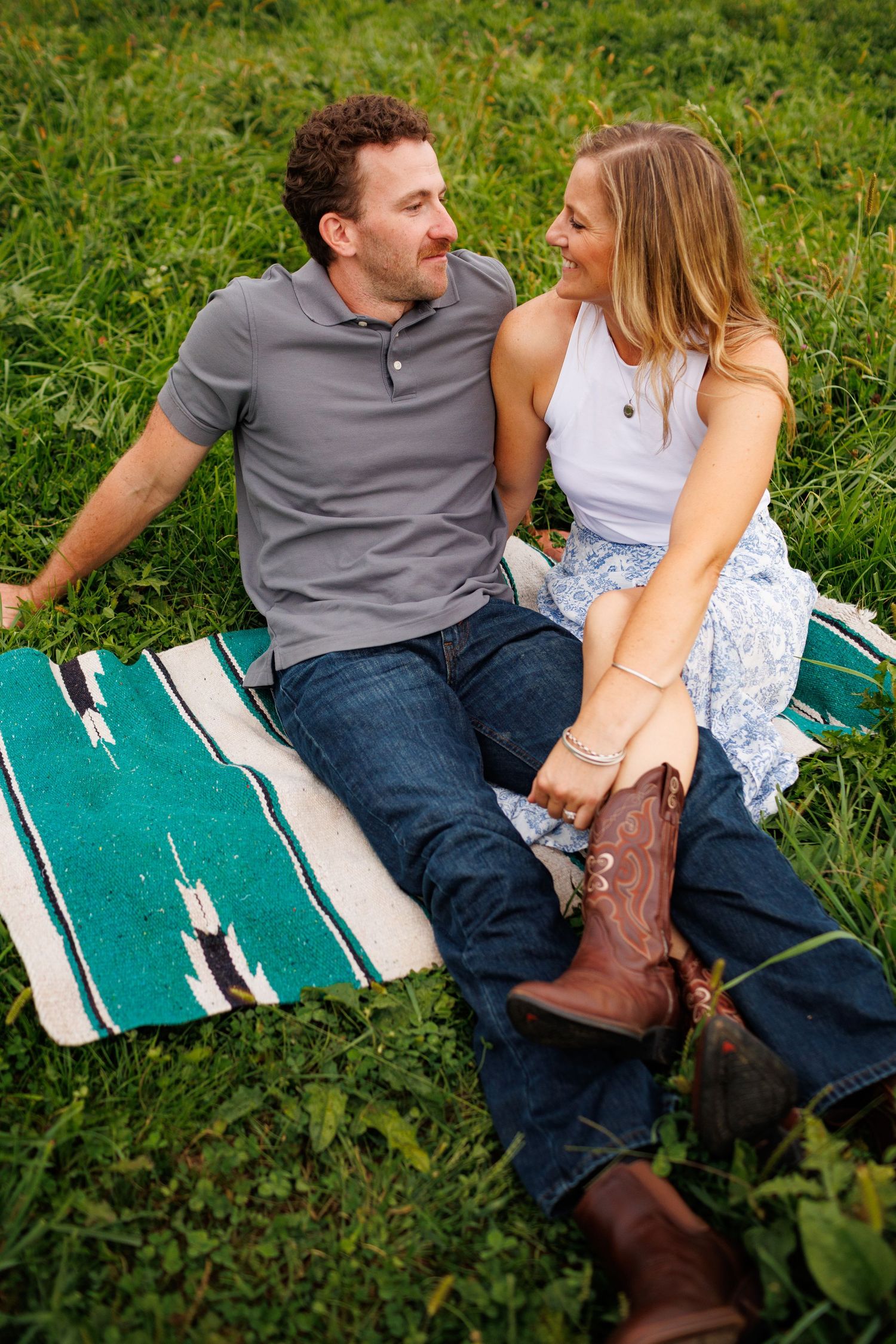 Couple sitting on a colorful blanket in green grass, smiling at each other during an outdoor engagement photo session.
