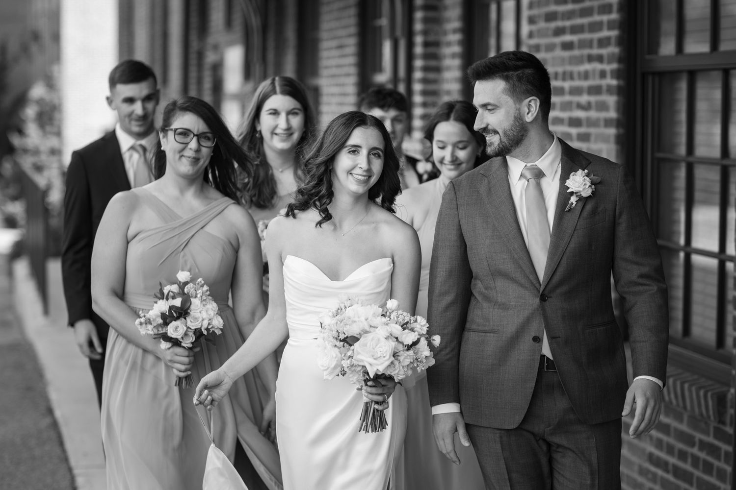 Black and white photo of bride and groom walking with wedding party past brick building.
