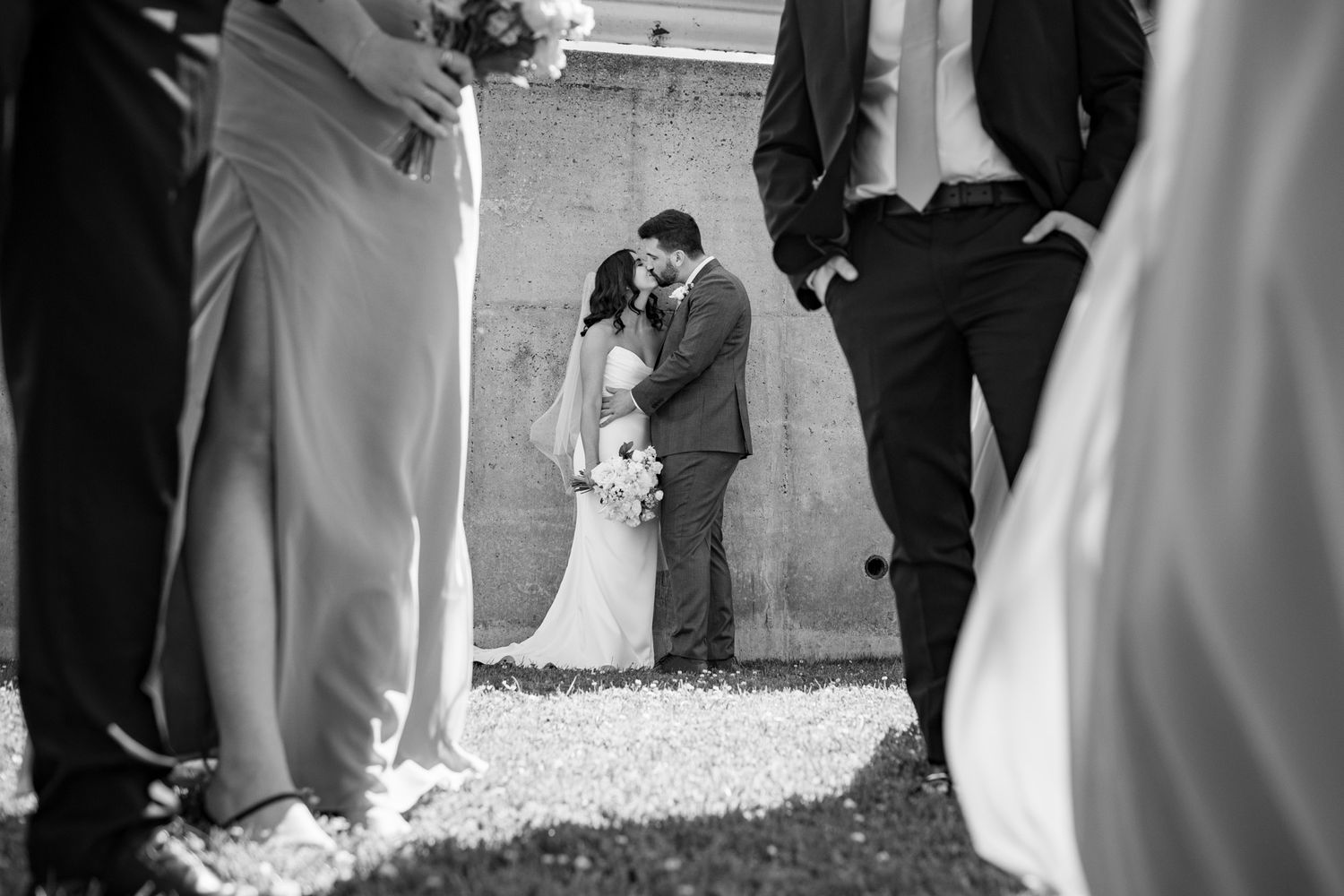 Black and white wedding photo of bride and groom kissing surrounded by wedding party outdoors.